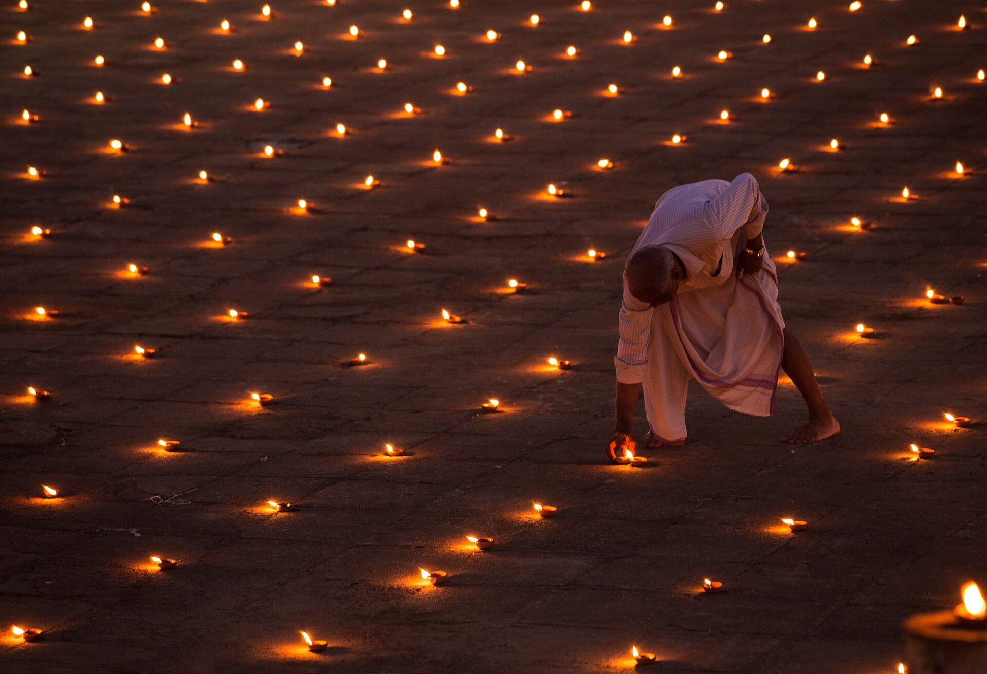 A man lits numerous candles lined in rows on the ground.