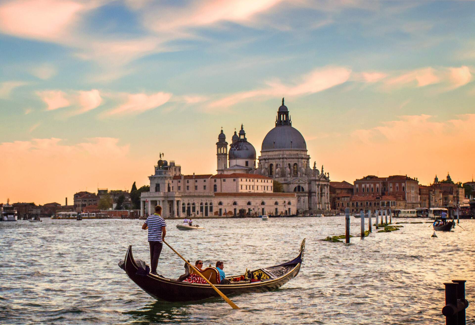 A man navigating a gondola with tourists across a water canal with distant views of a dome church under an orange sky.