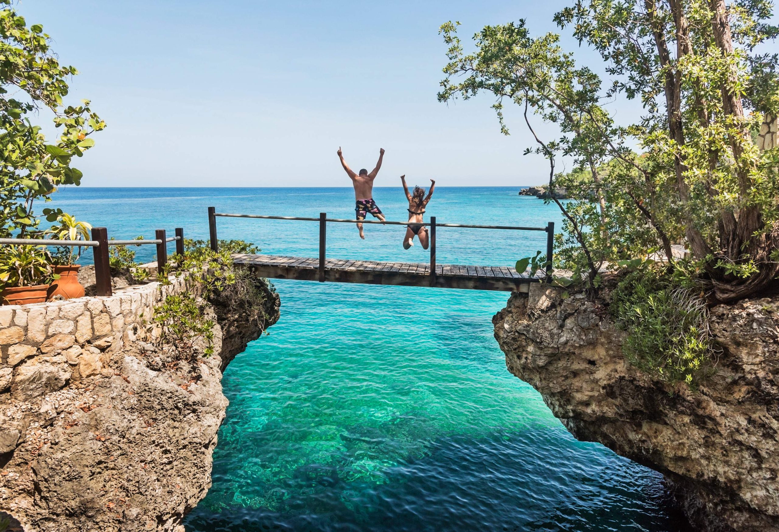 Two people jumping off a short bridge into the turquoise ocean.