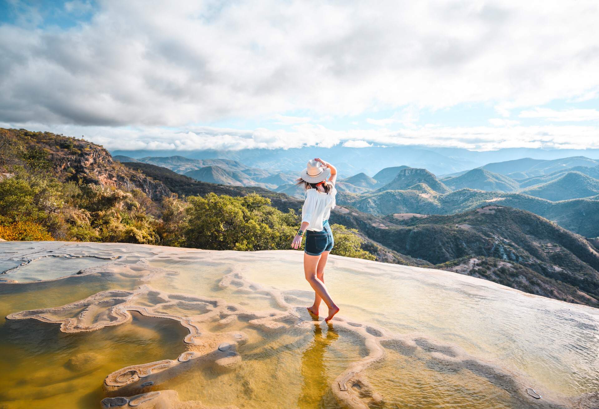 A woman stands on the natural rock formations where water trickles over the cliff overlooking the hills and cloud-capped mountains IN HIERVE EL AGUA