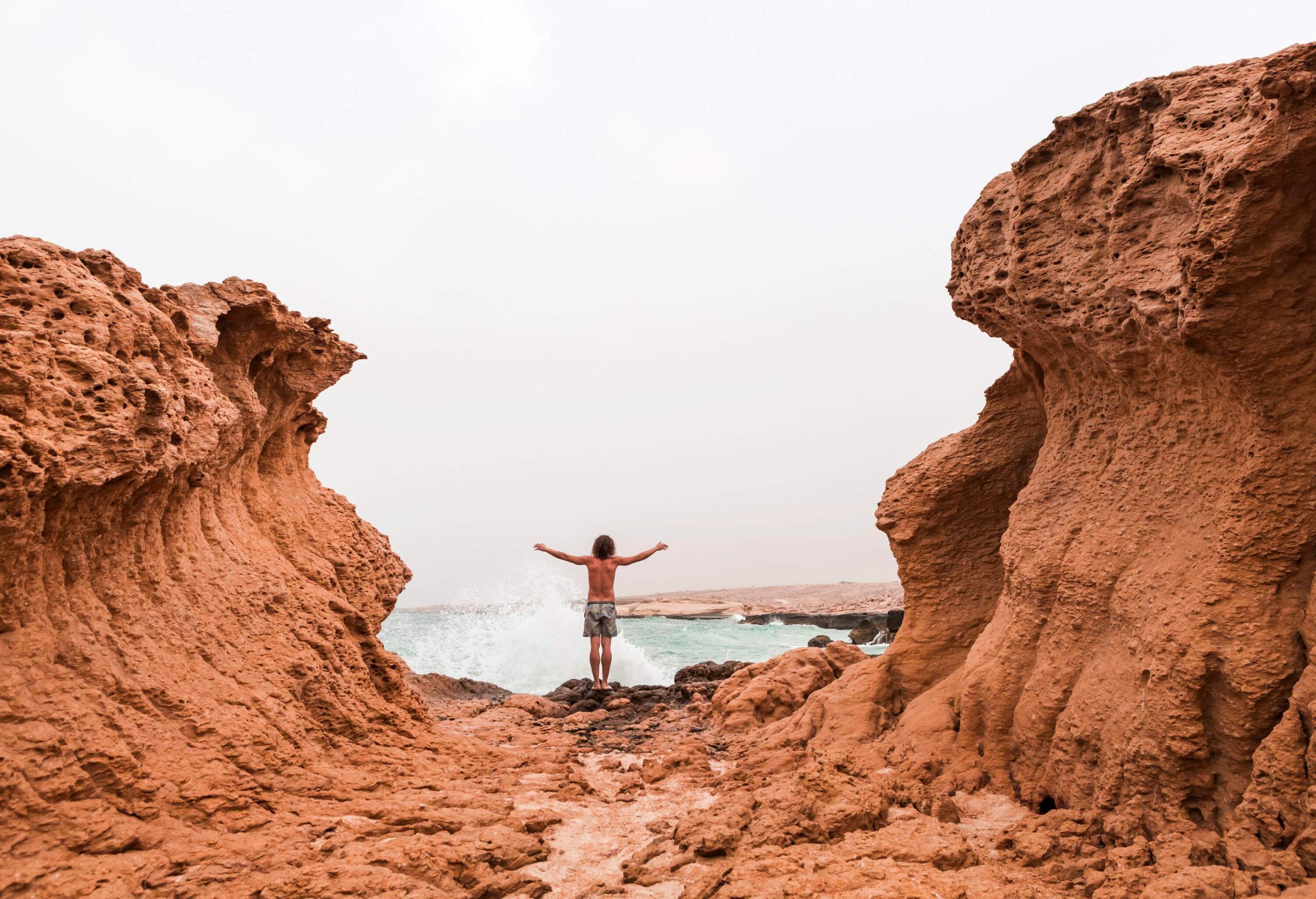 A rear view of a man with his hands raised to the side while standing on the rocks alongside the sea between two rock formations.