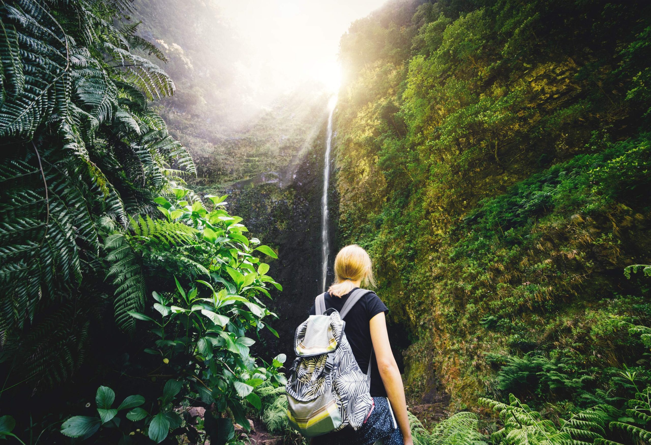 A blonde woman with a backpack gazing up at a tall, sunlit cascade.