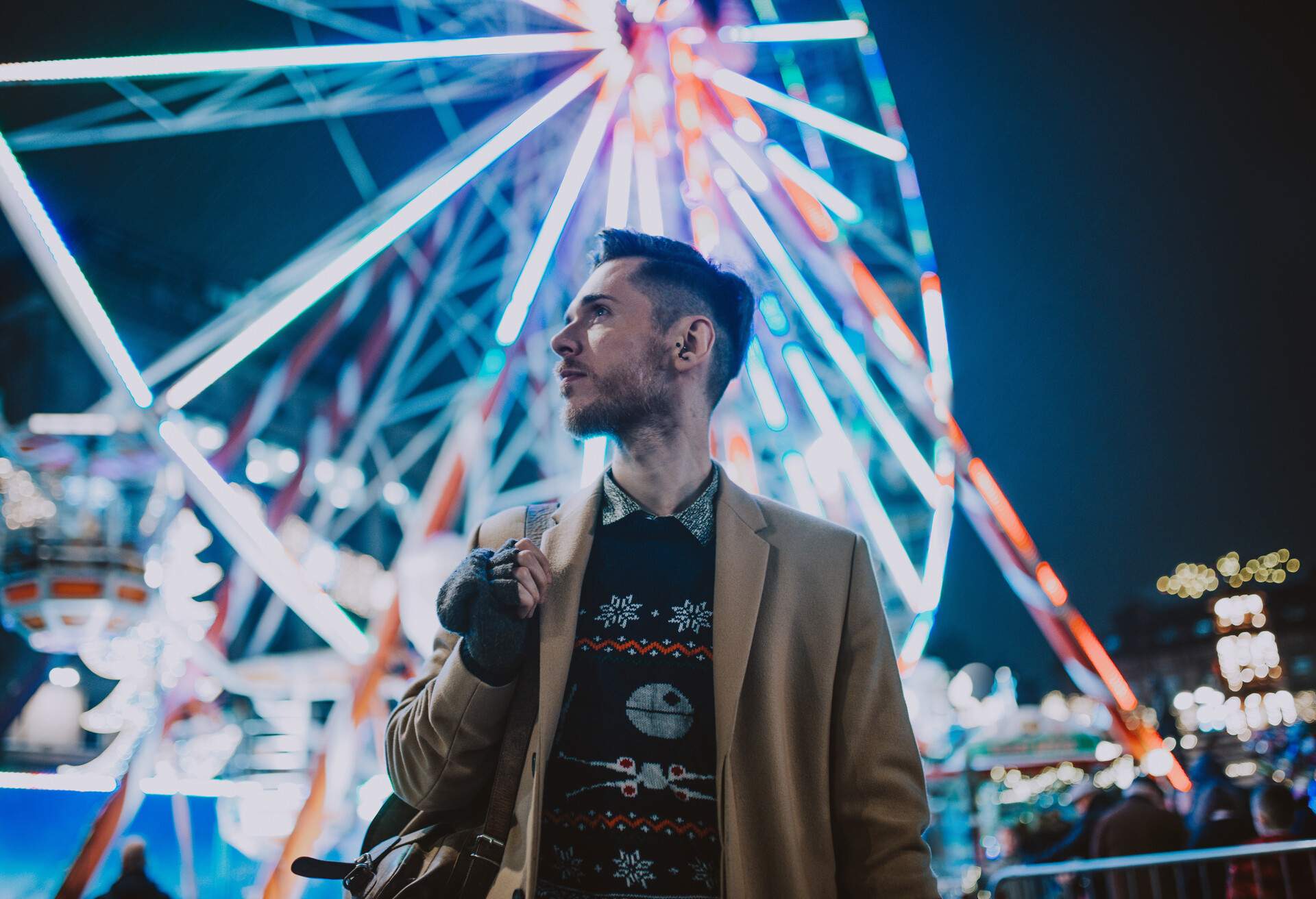 A man in warm clothing stands against a backdrop of a Ferris wheel with bright lights.