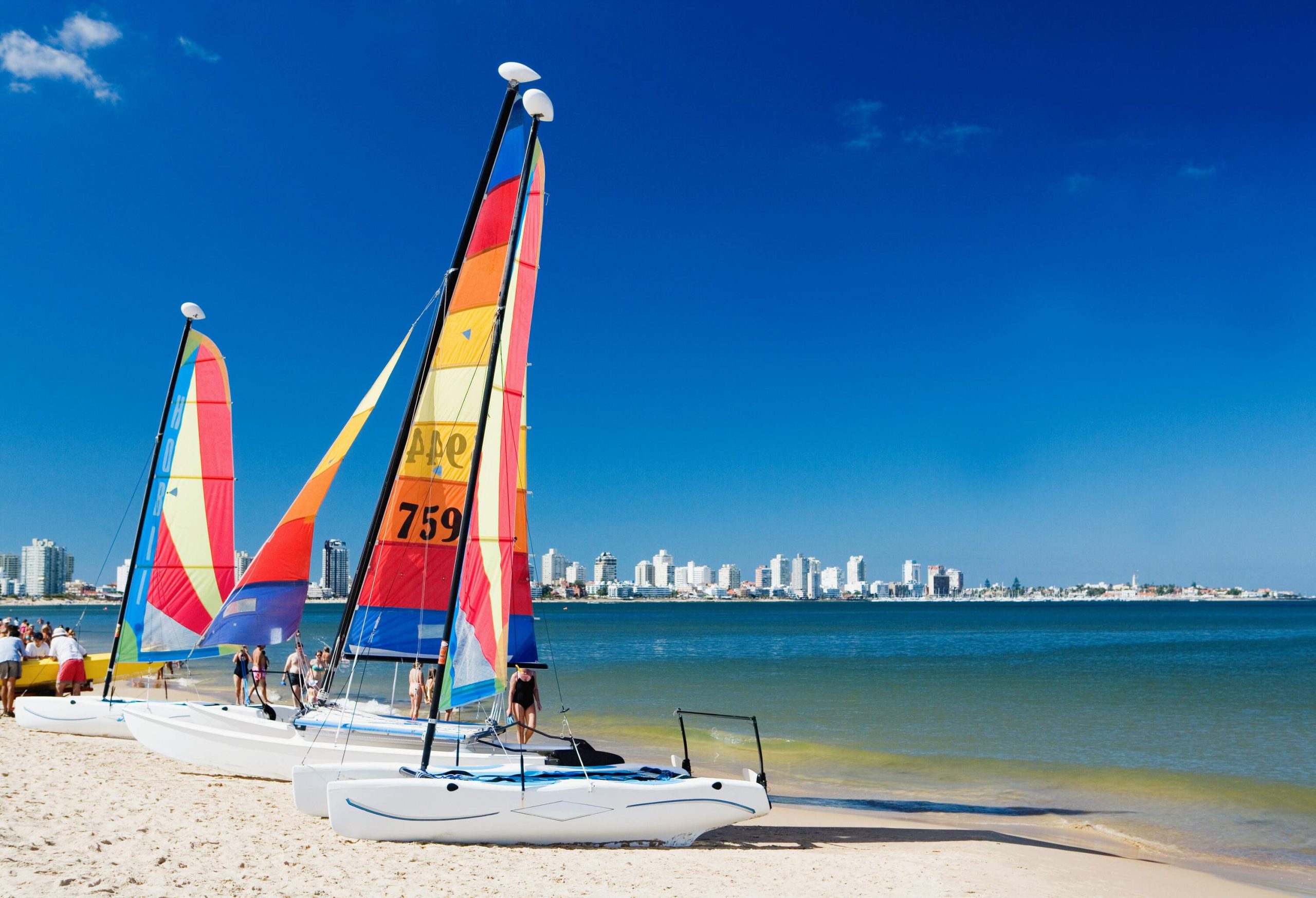 Colourful sailboats docked on the fine white sands, with some tourist passing by and a view of a cityscape in the distance.