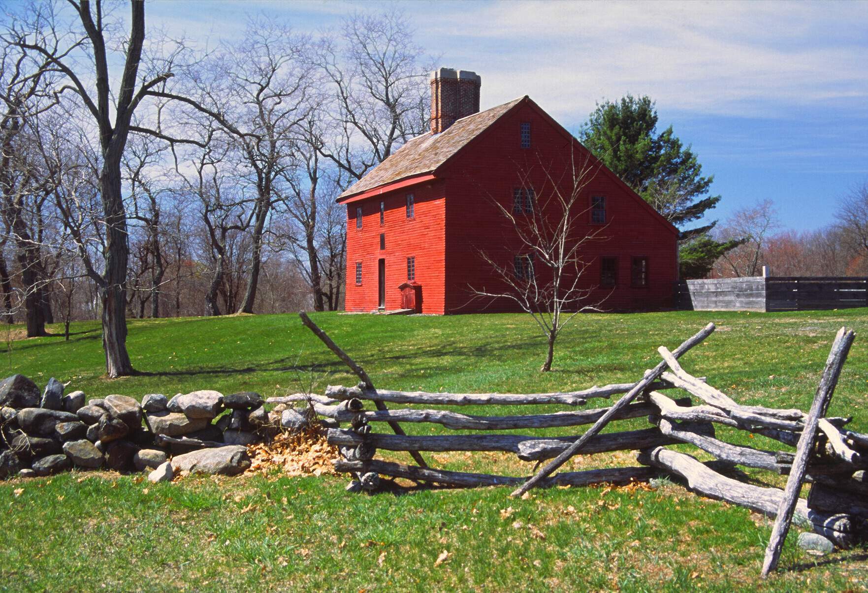 A brick house with a raised chimney on a green lawn amongst bare trees enclosed in stone walls with wooden gates.