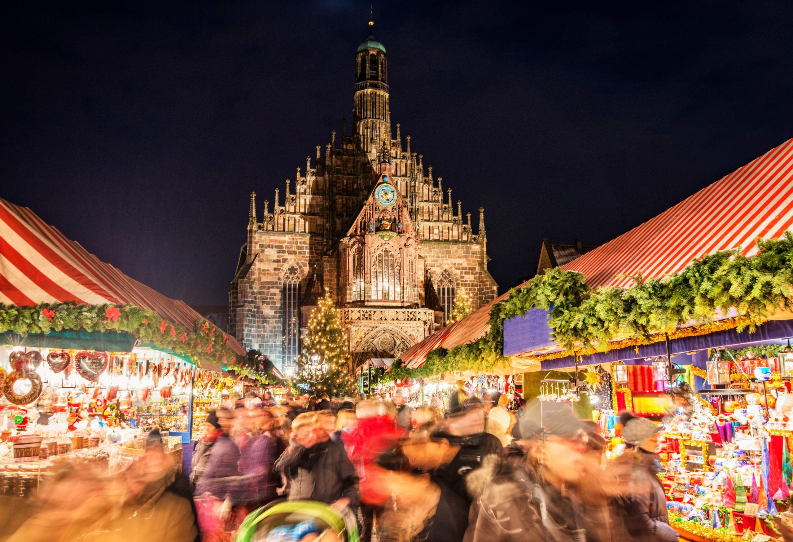 A brick Gothic church behind a crowd of people in motion, in a busy Christmas market at night with colourful illuminated Christmas decorations and food stalls. 