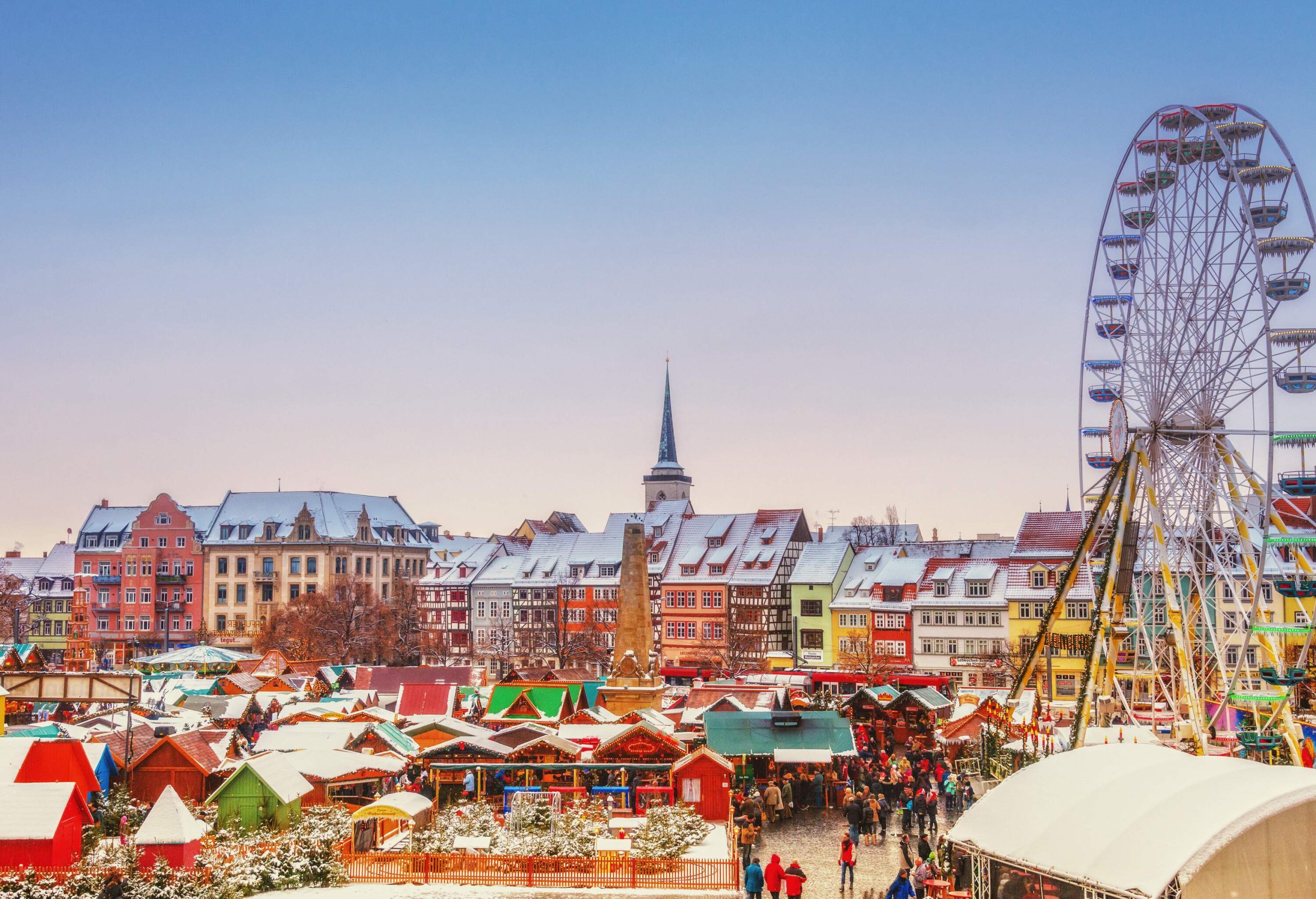 A packed Christmas market with vibrant stalls, a Ferris wheel, and colourful buildings in the backdrop.
