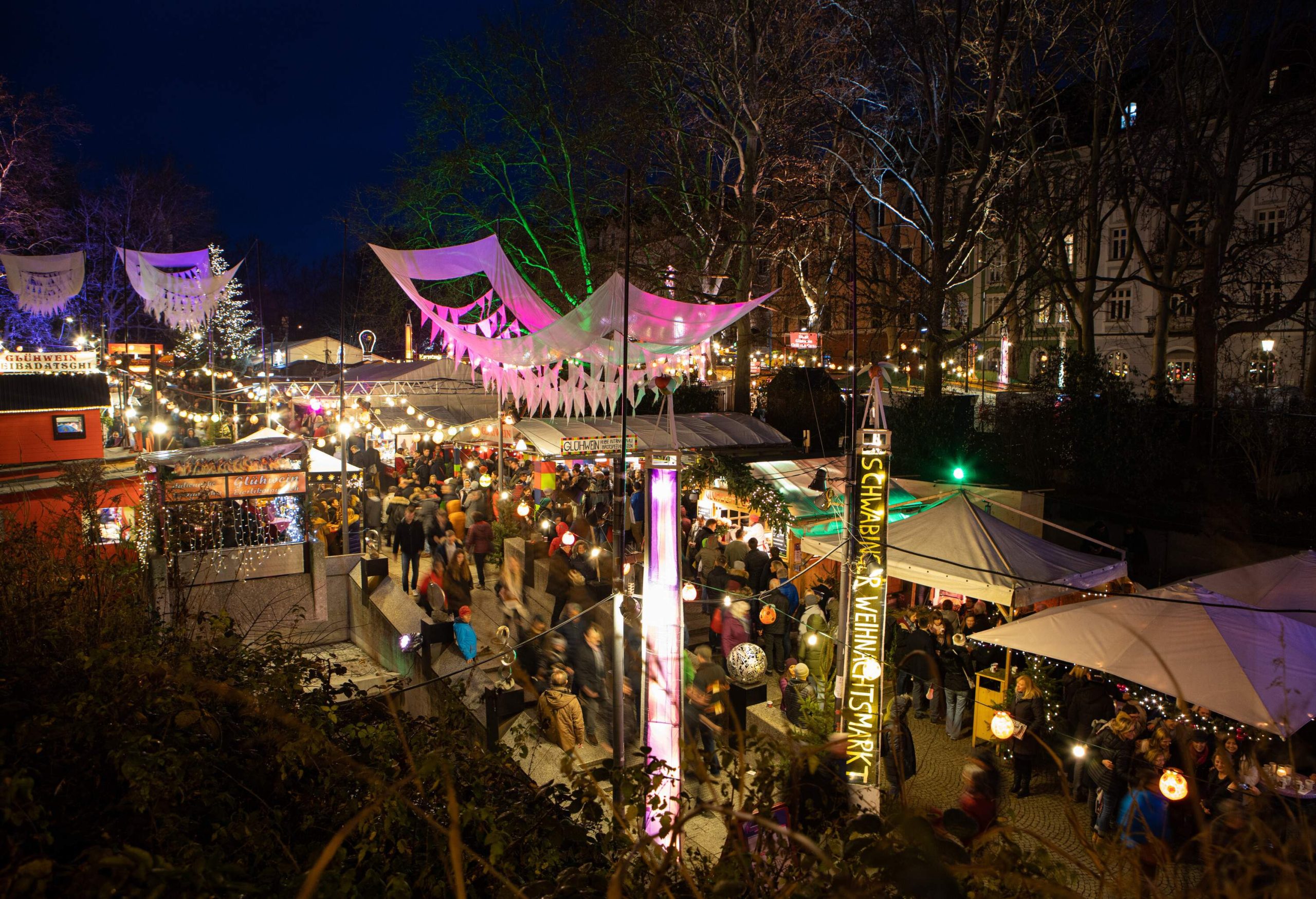 People are in motion in the brightly illuminated Christmas market lined with tall bare trees.