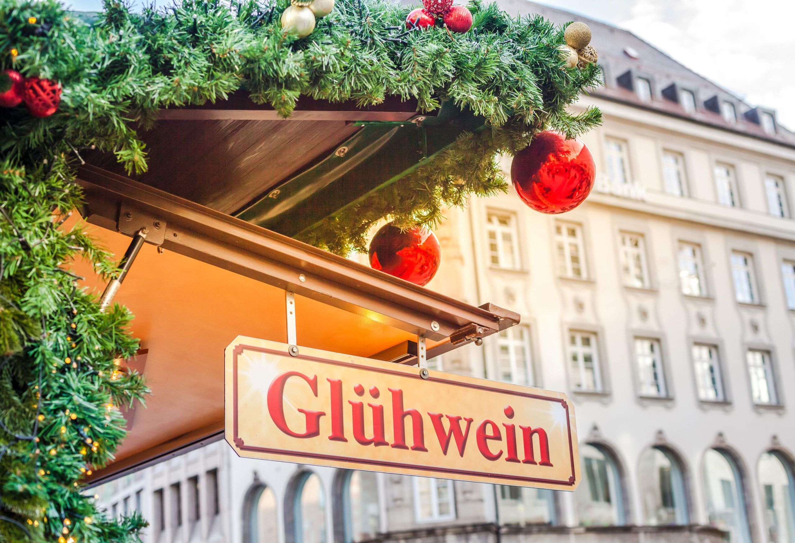 A gold-plated sign board reading "Glühwein" attached to the roof decorated with Christmas decorations and a view of an old building in the background.