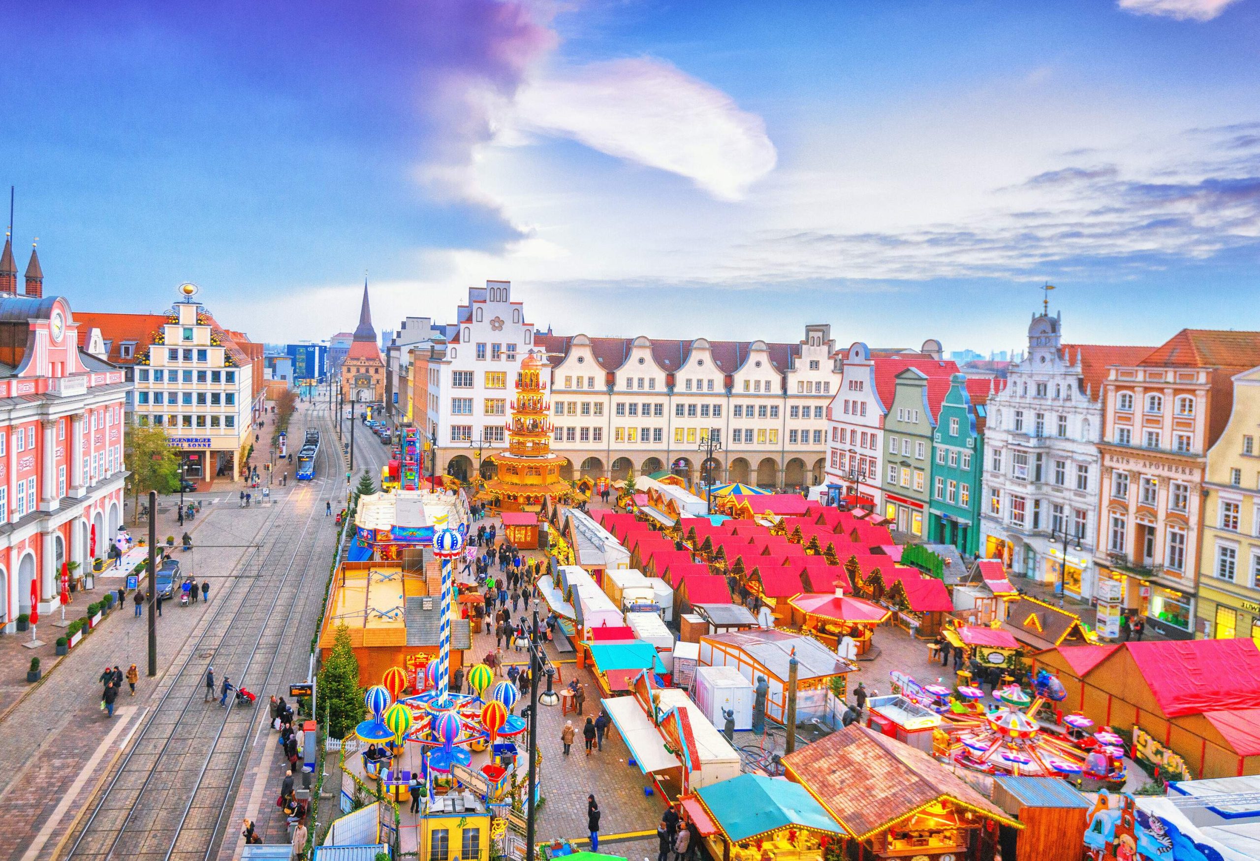 A town square, surrounded by gabled houses of different colours, with a train line on one side and a vibrant Christmas market on the other.