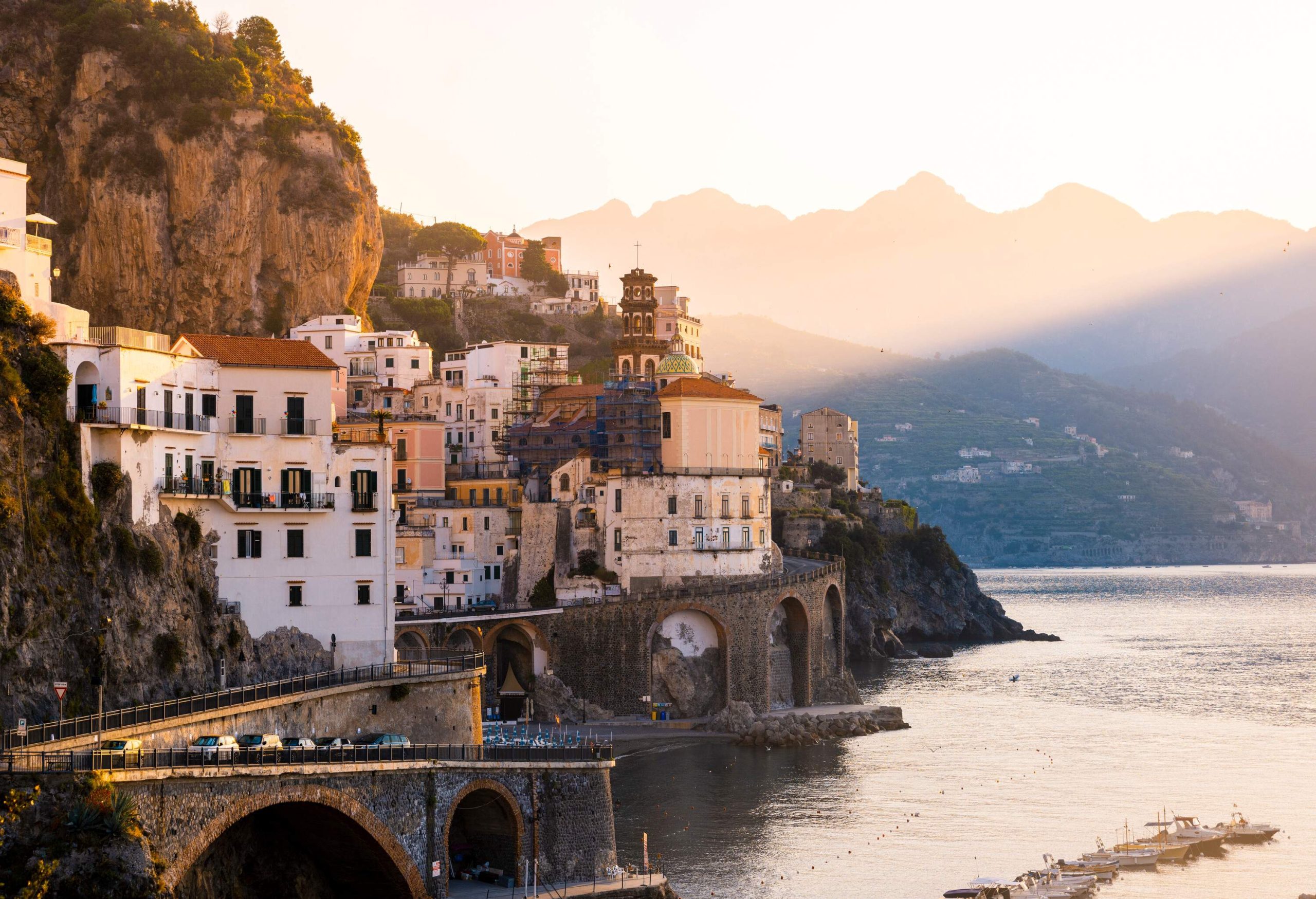 A sunlit coastal village on the steep mountainside overlooking the lined small boats on the sea.