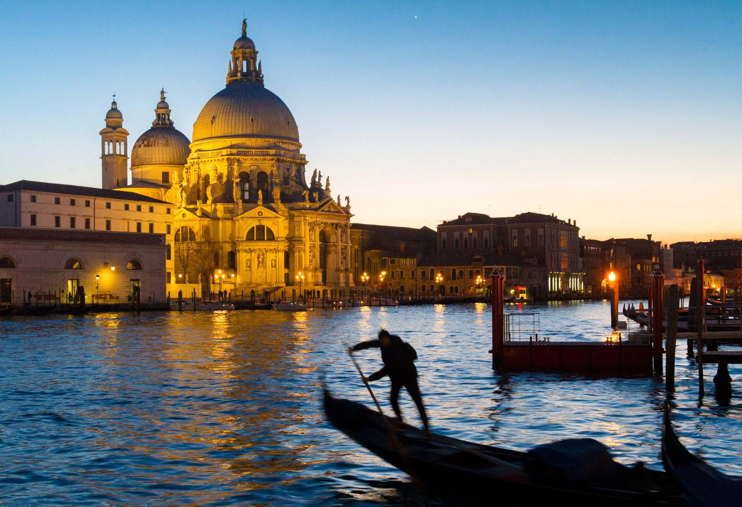 Silhouetted gondolier in the Grand Canal at Punta della Dogana with illuminated St Mary of Health (Santa Maria della Salute) Basilica at dusk. Venice, Veneto, Italy.