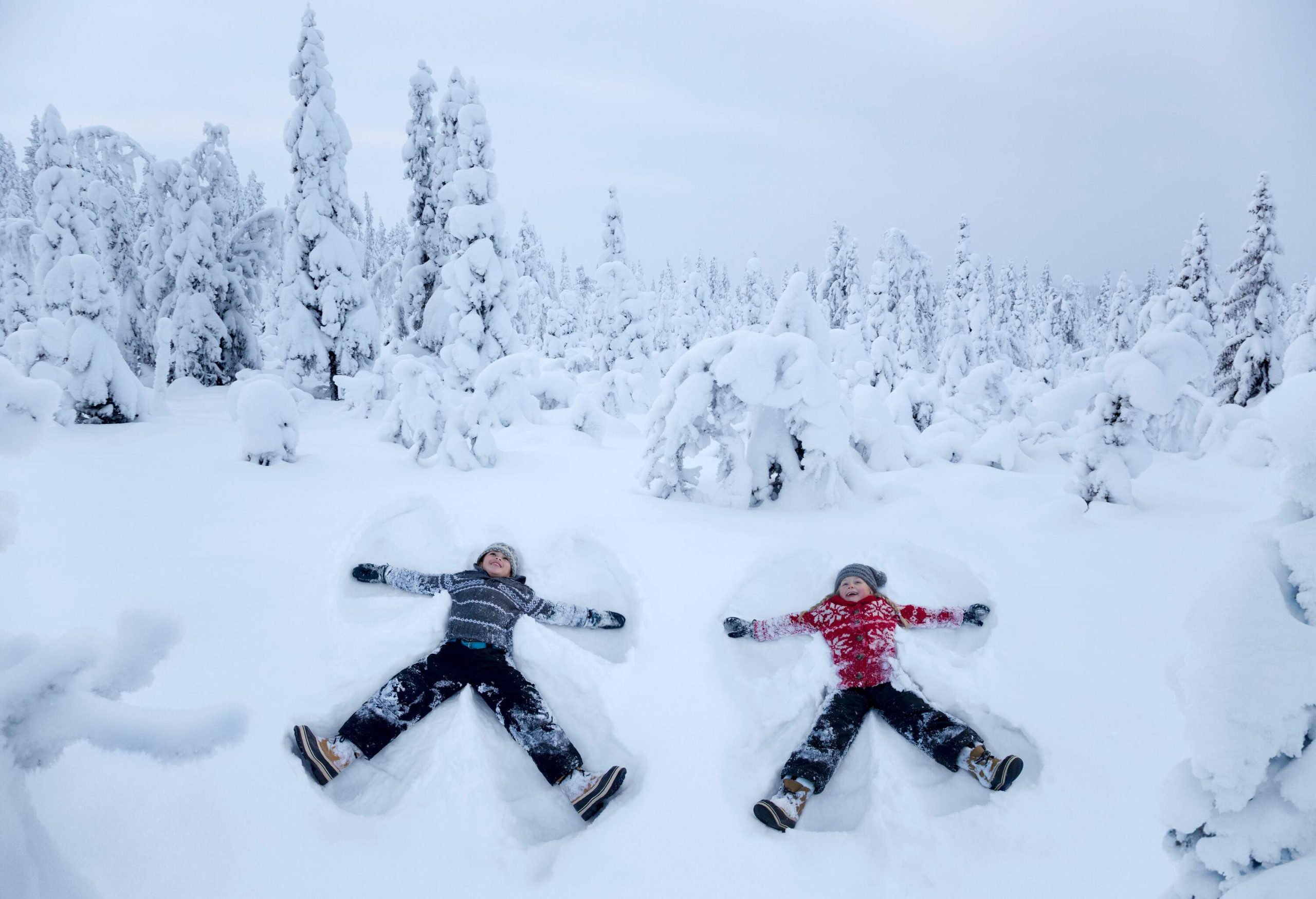 Two kids making snow angels in the snow, with heavily frosted trees behind them.