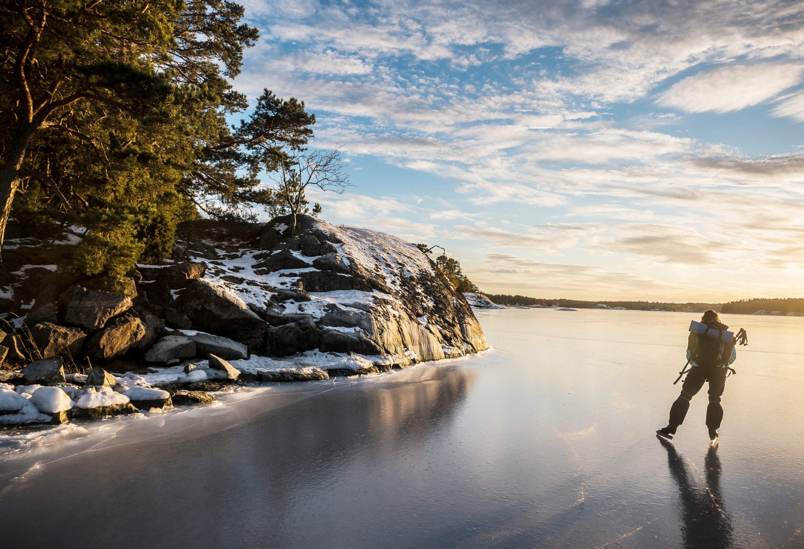 A person skates on a frozen lake beside the boulders and trees while carrying a camping gear.