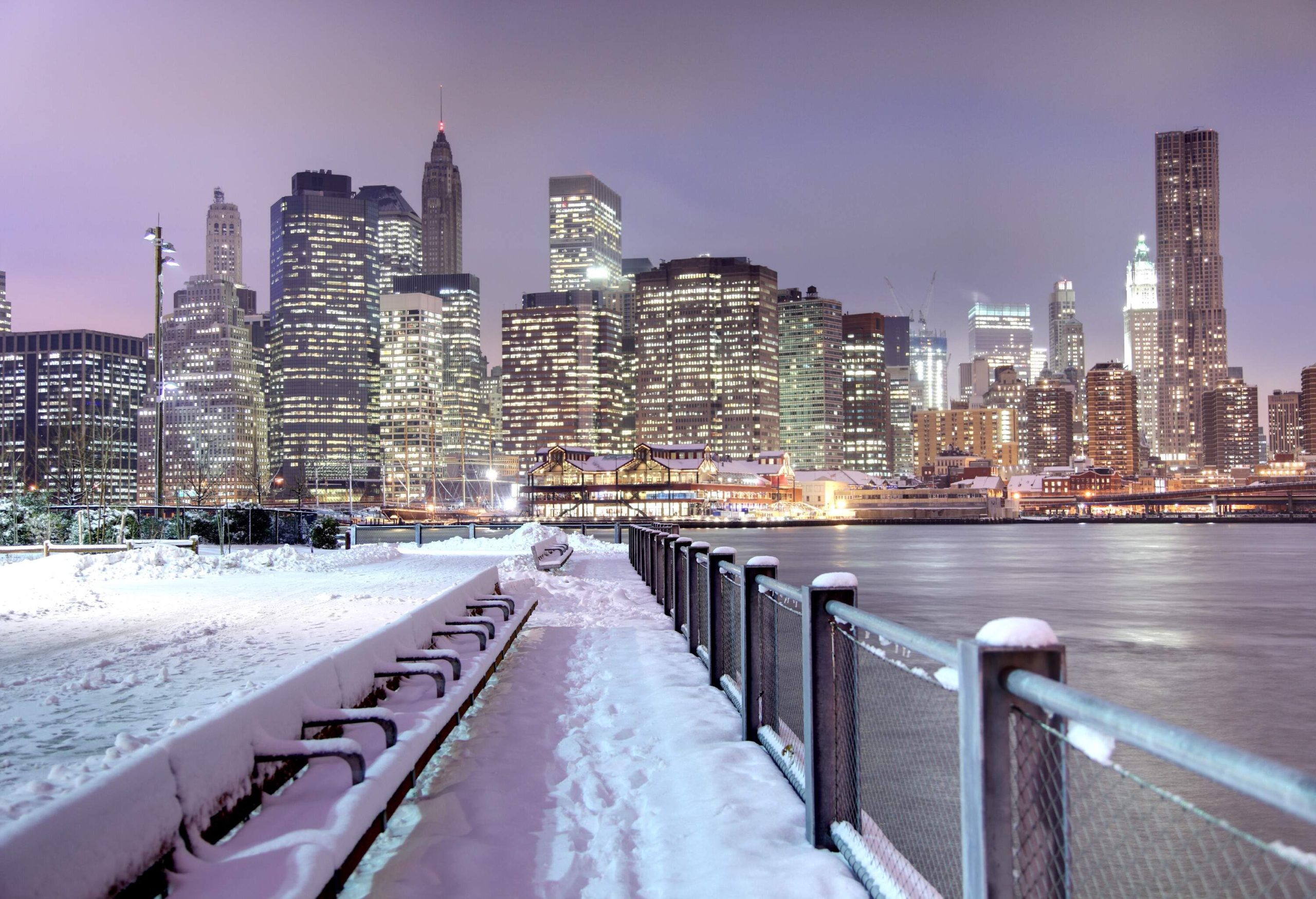 Snow-covered benches on a fenced riverside walkway with views of New York City's illuminated skyline.