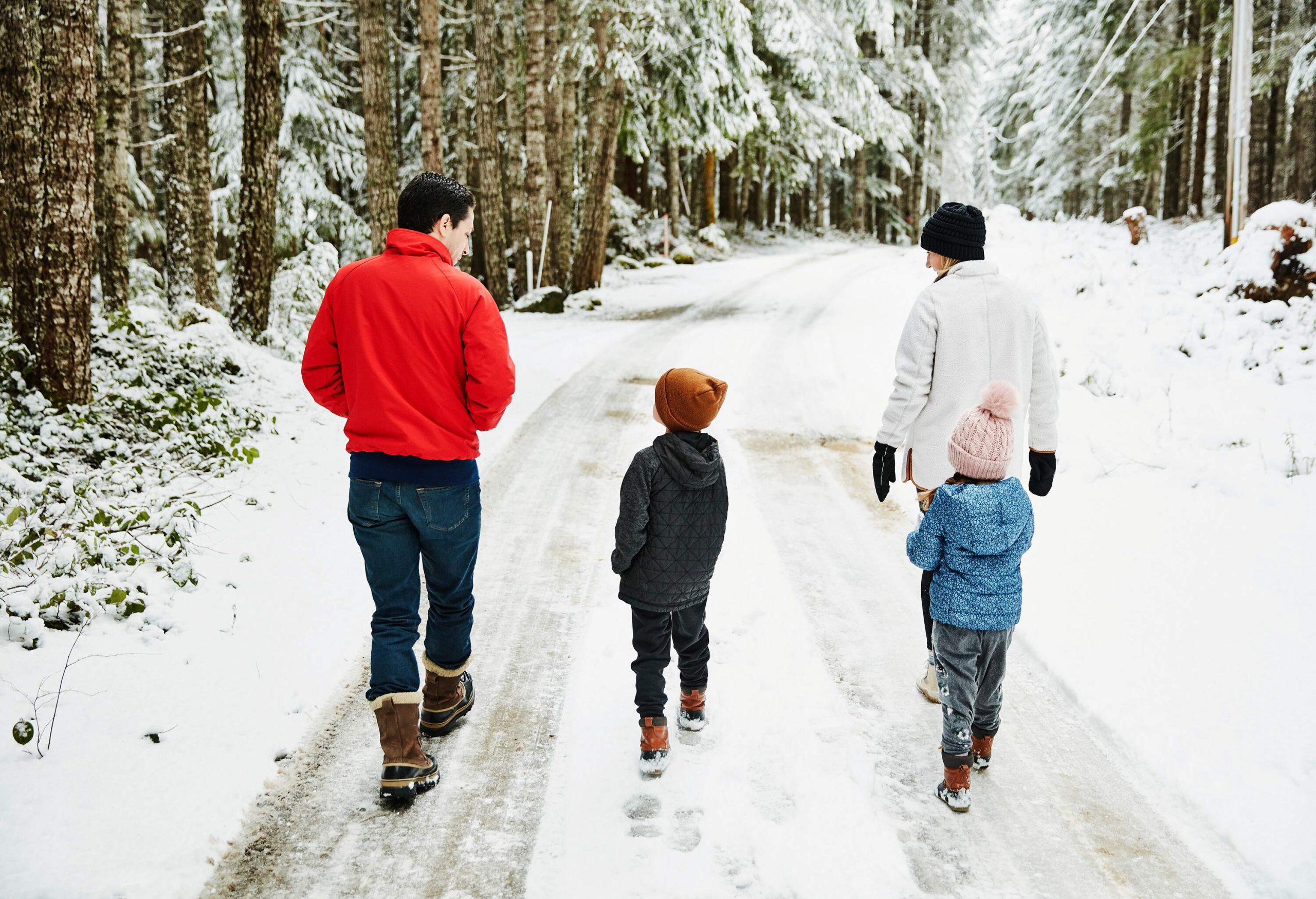A family of four in winter attire is walking along a snowy road lined with tall frost-covered trees.