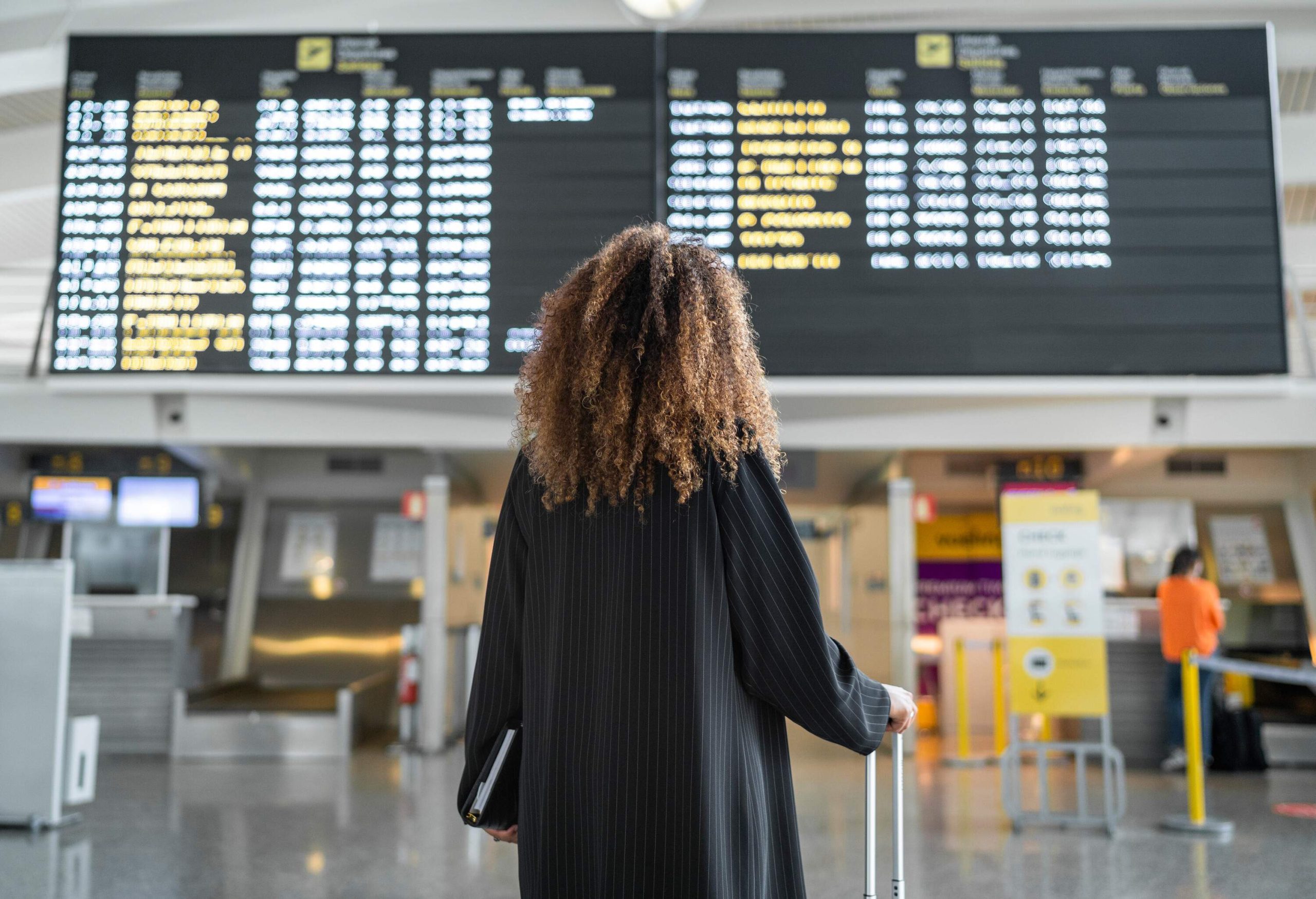 A curly-haired person in a black dress stands in front of an airport flight information display.