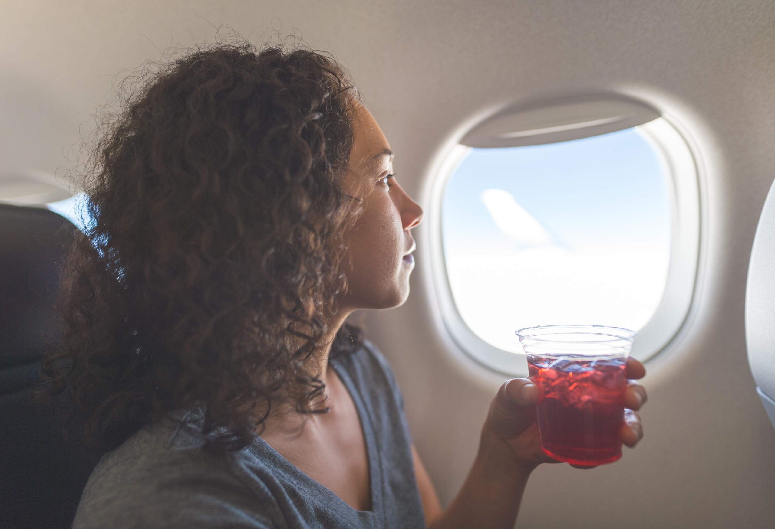a young woman with curly hair looking out the plane window while holding a drink in her hand.