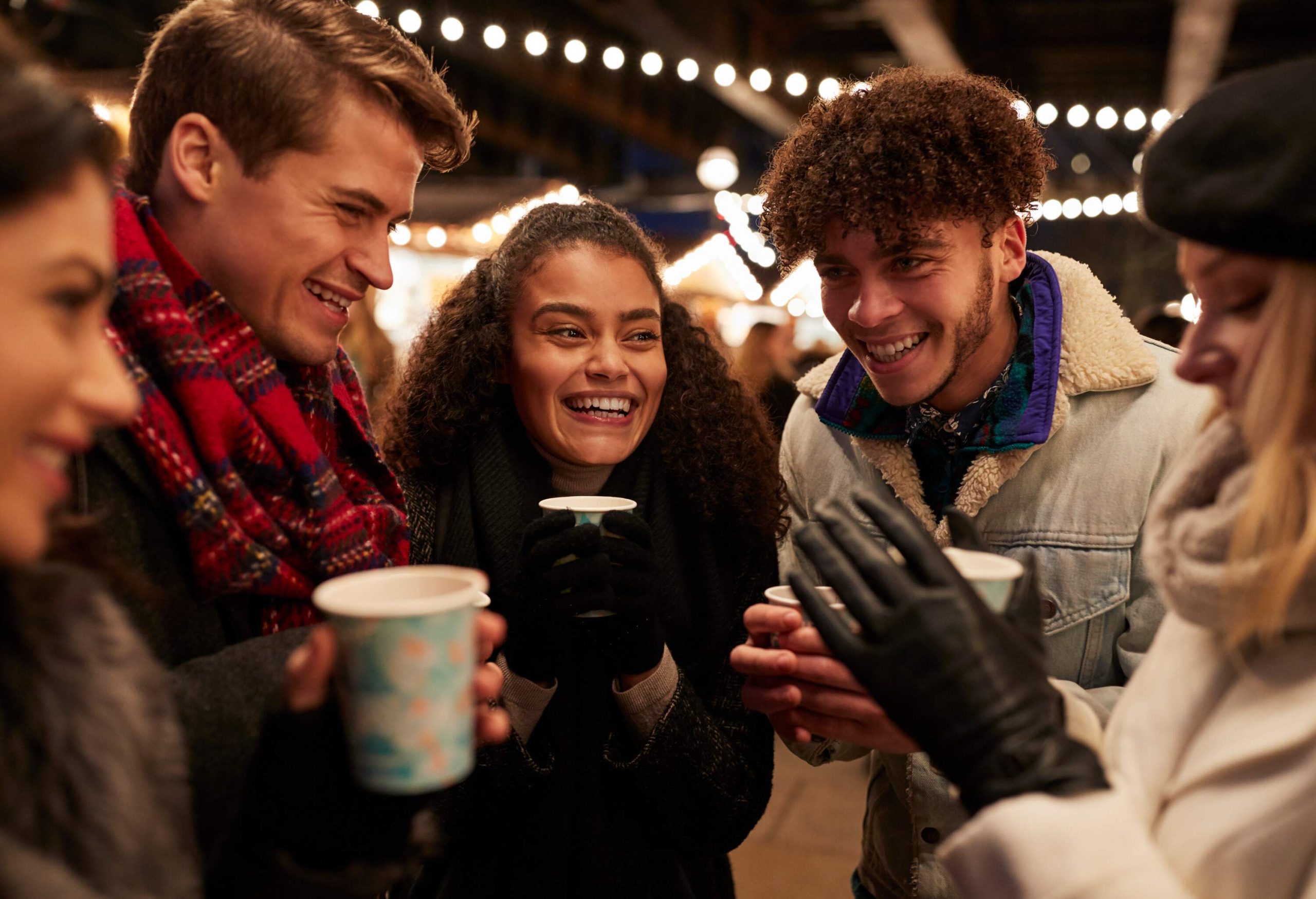 Five friends in warm clothing face each other while smiling and holding their cup of drinks.
