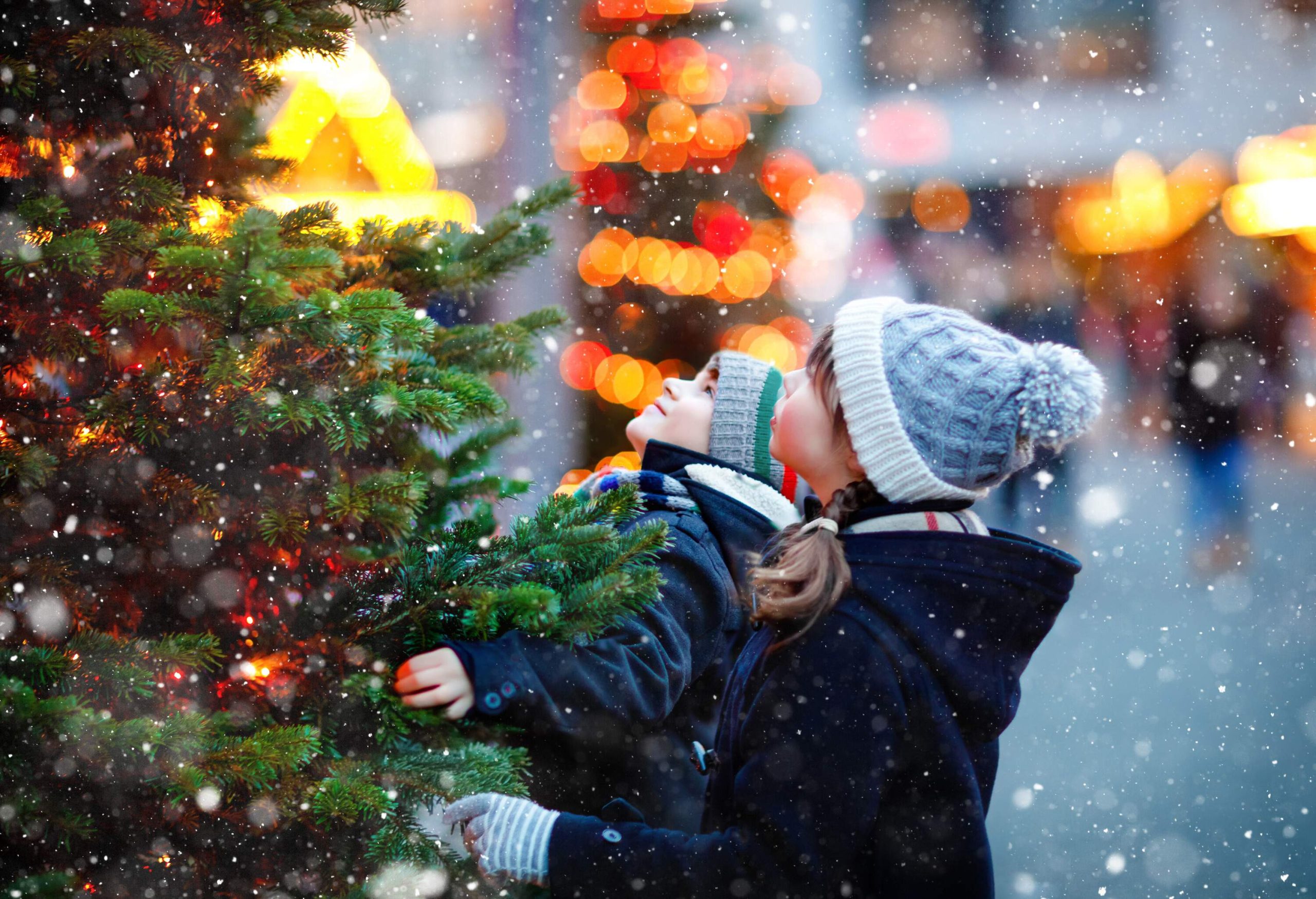 Two kids in winter jacket and knitted cap gaze up to a Christmas tree during a snowfall.