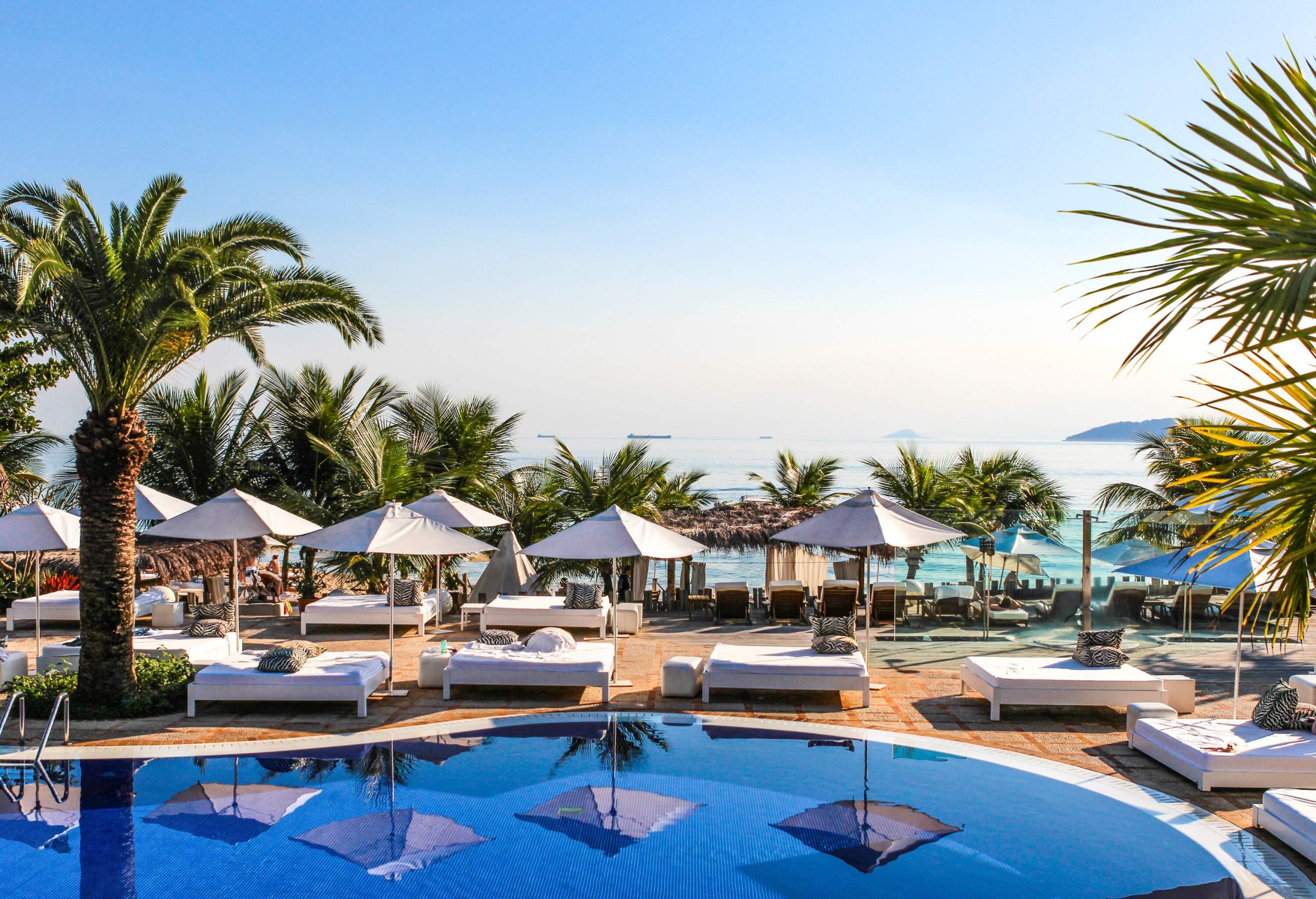 A swimming pool area, surrounded by foam-padded sun beds under parasols, extending to another row of sun beds lined up facing the sea, all framed by a tranquil backdrop of palm trees.