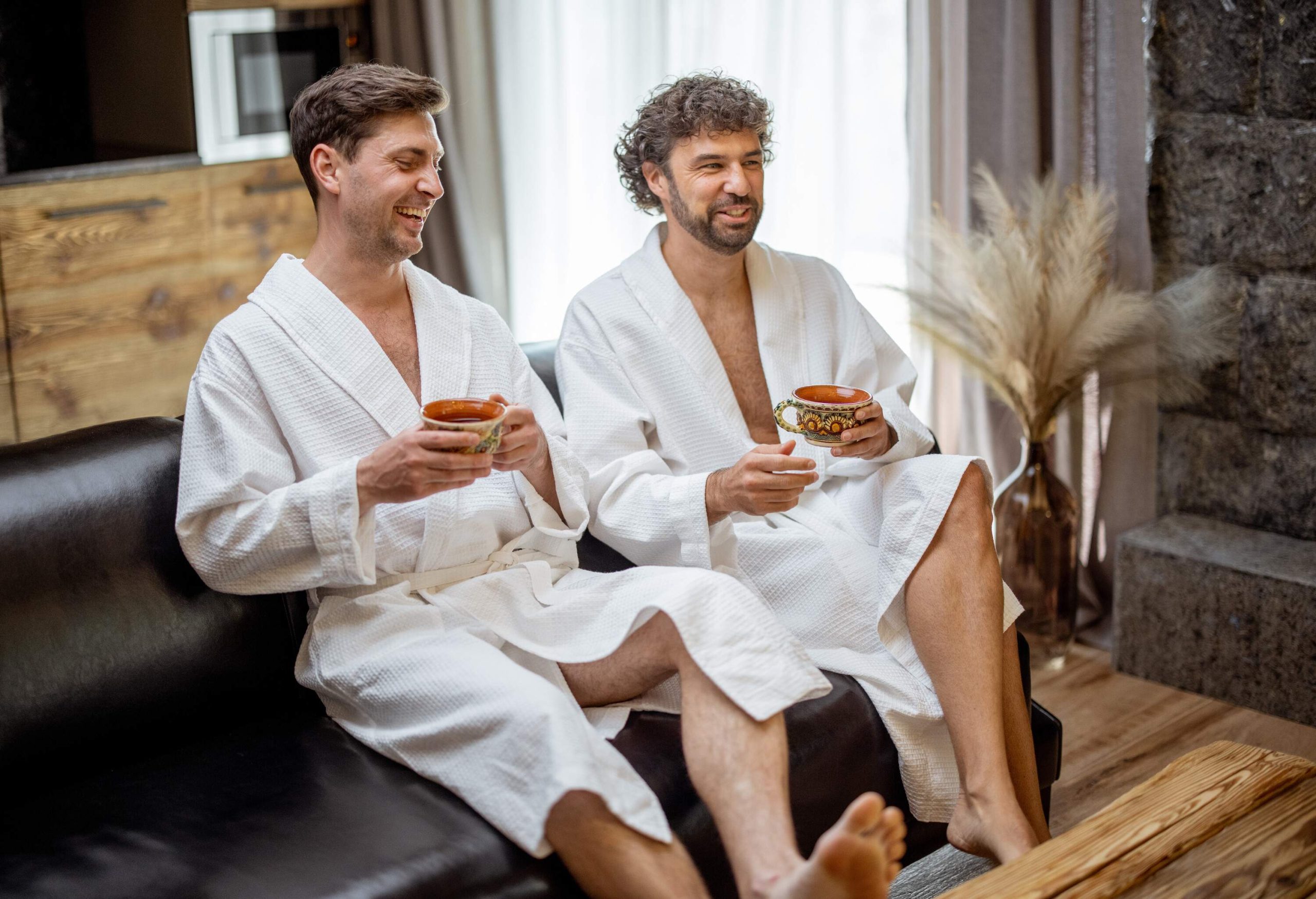 Two men wearing white bathrobes sitting on a leather couch, drinking coffee.