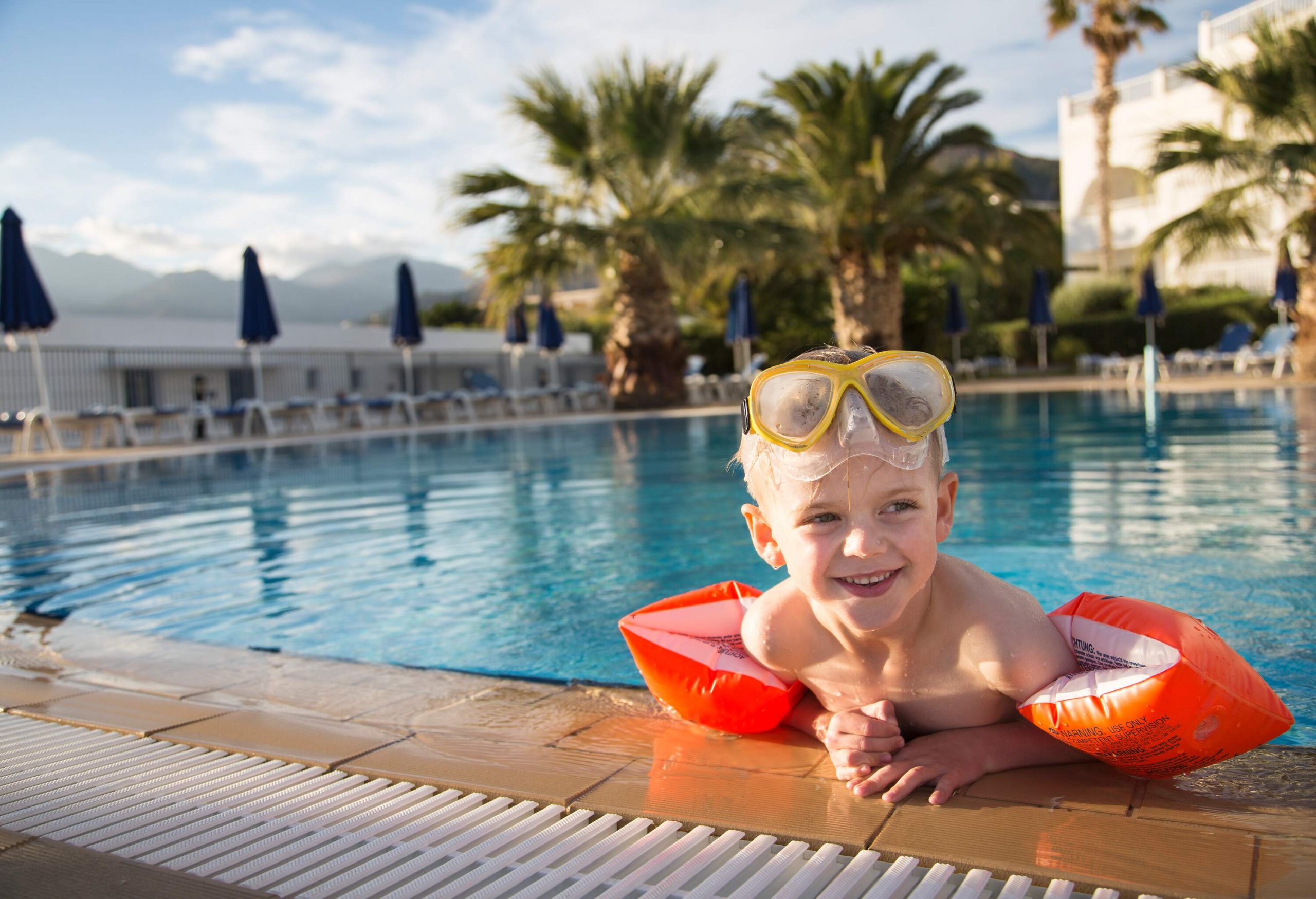 A happy little boy wearing a pair of goggles and arm floaties resting on the pool's edge.