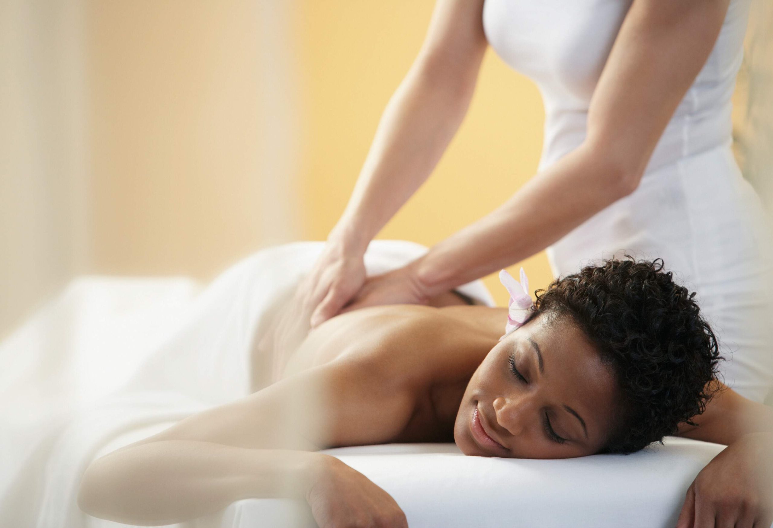 A woman lies on a spa bed with her eyes closed while receiving a back massage.