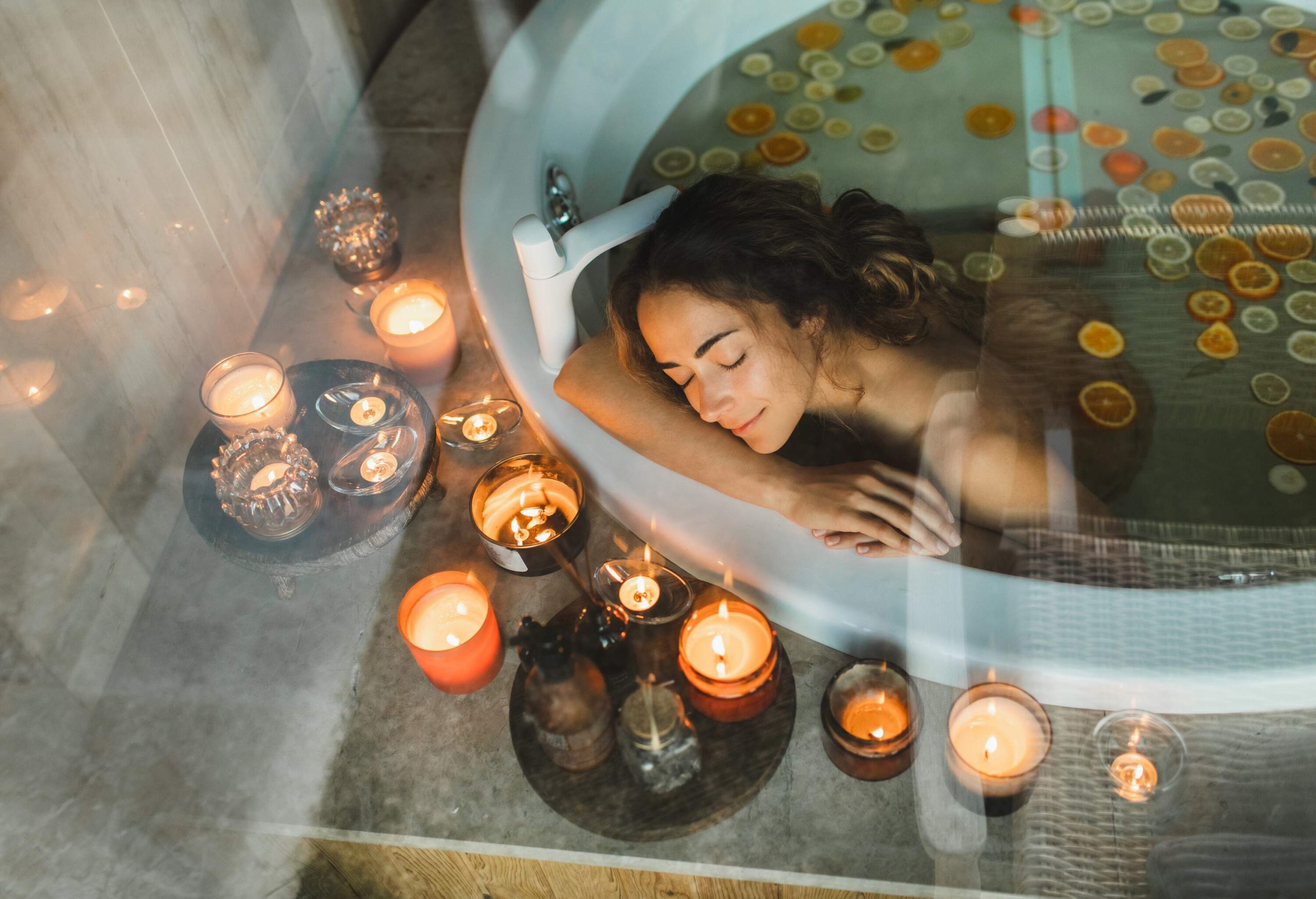 Relaxing scene of a beautiful woman sitting in a bath filled with slices of fruit, surrounded by softly lit candles.