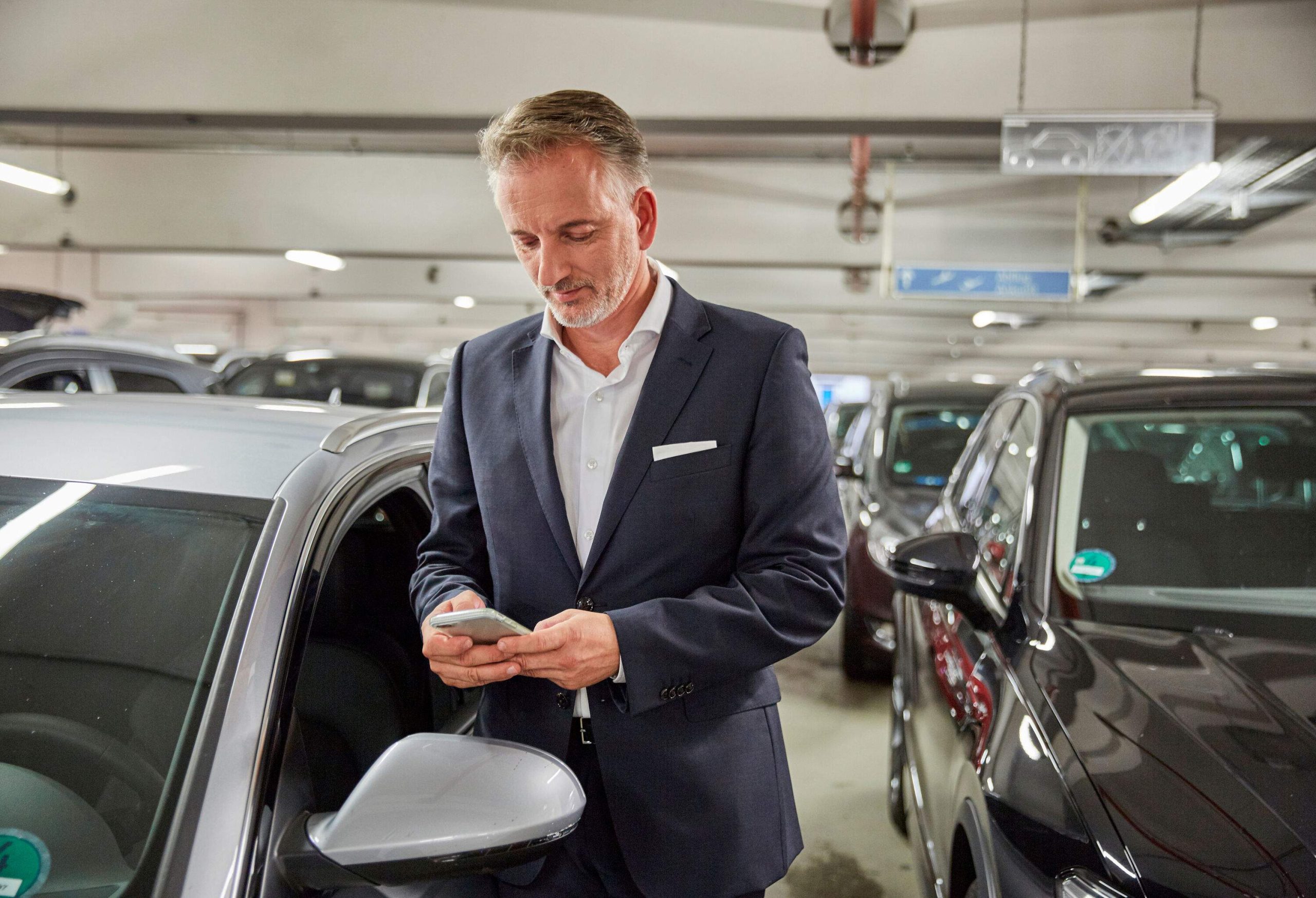 A person dressed in corporate attire stands beside a pristine white car, engrossed in their smartphone, amidst the ambiance of an underground parking area.