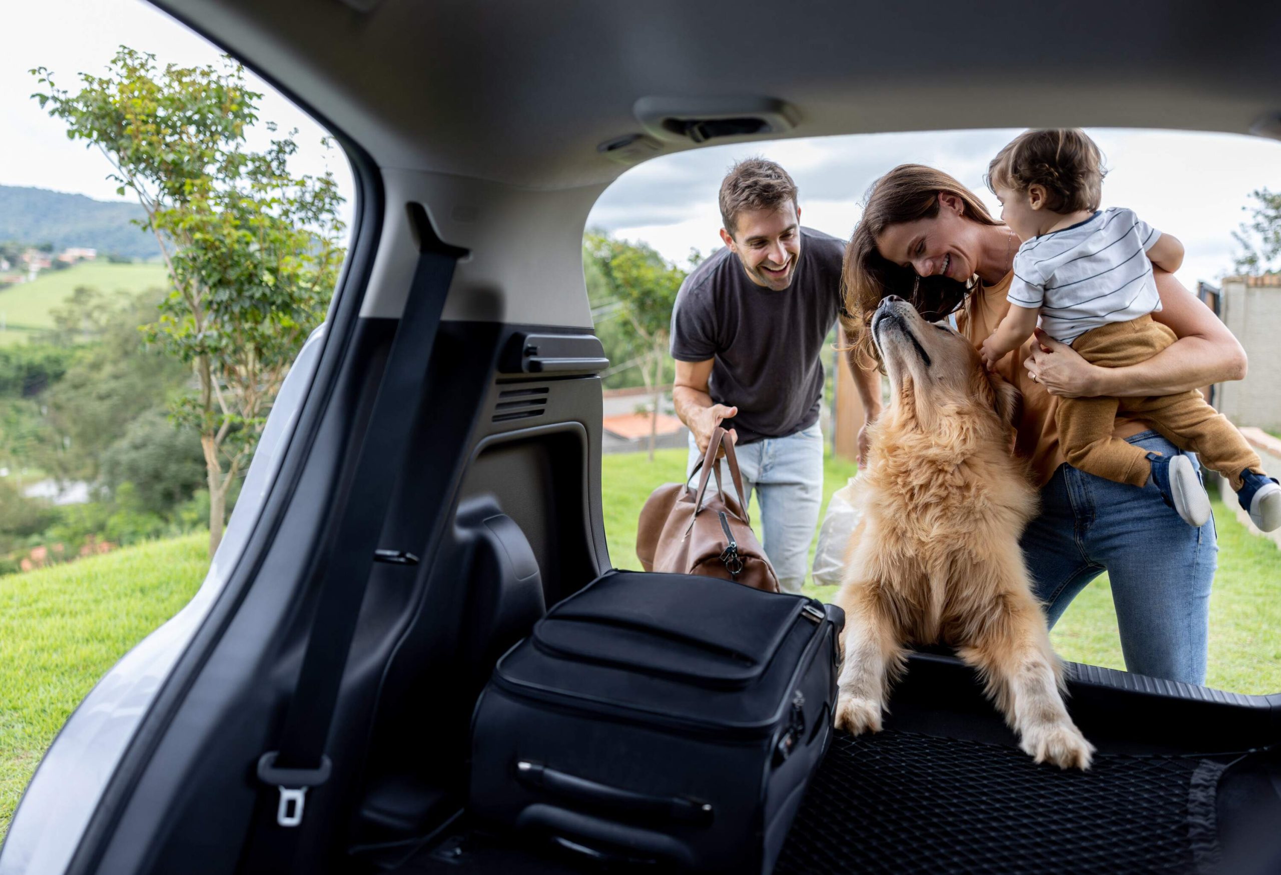 A woman holding a small child petting a large dog and a man loading bags into the car trunk.