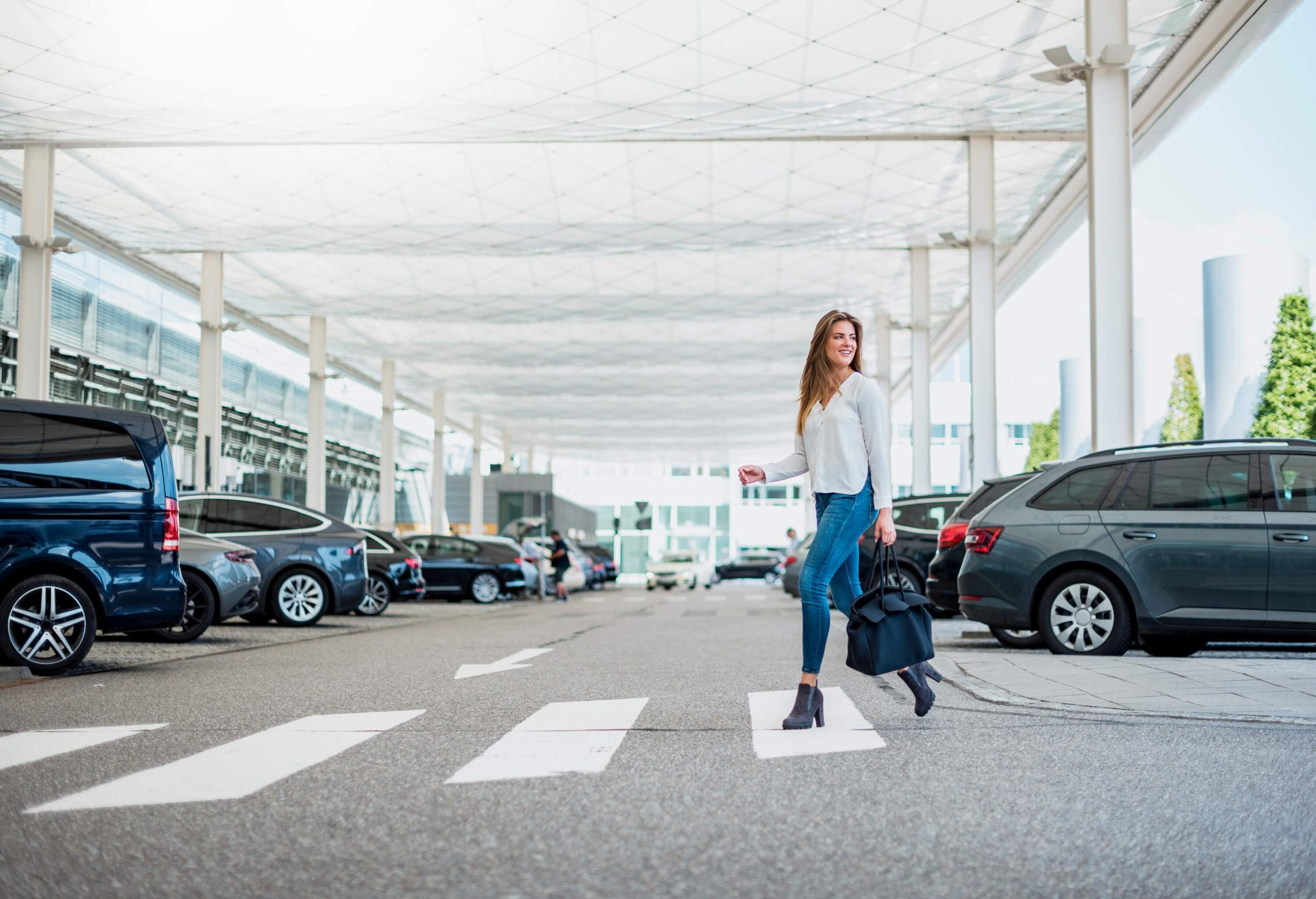 A woman in casual clothes carries a bag as she crosses a crosswalk in a parking lot.