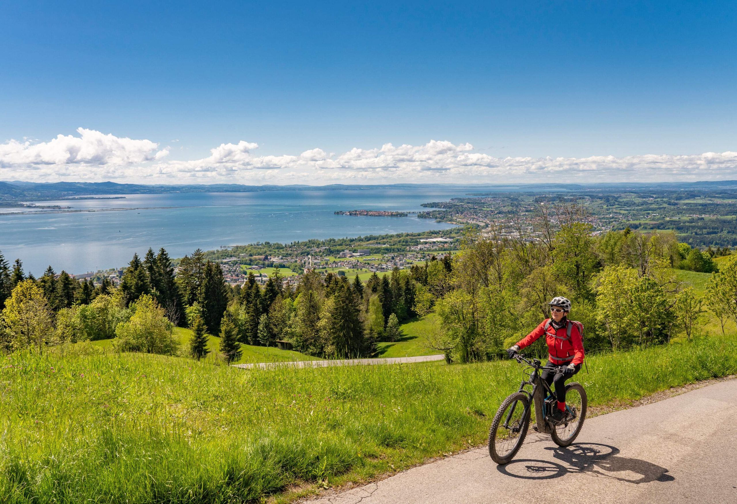 A woman on a bike cycling along a grass field and trees with a large lake in the distance.