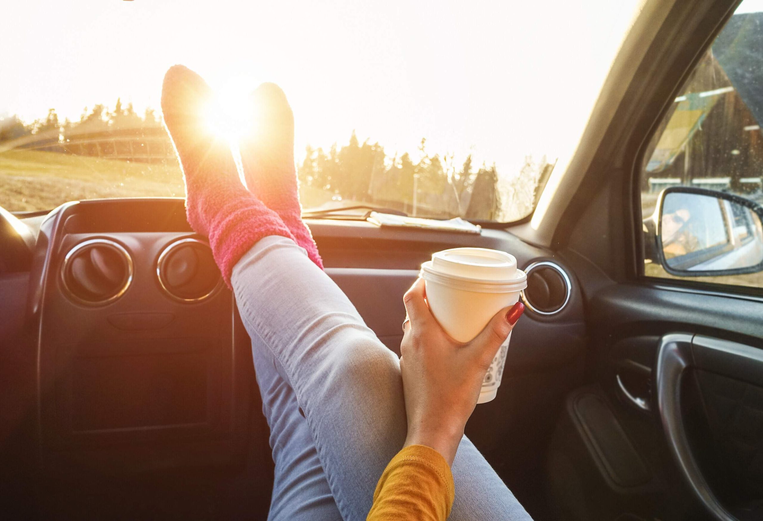 A woman sitting inside a car with a coffee cup in her hand, her sock-clad feet resting on the dashboard.