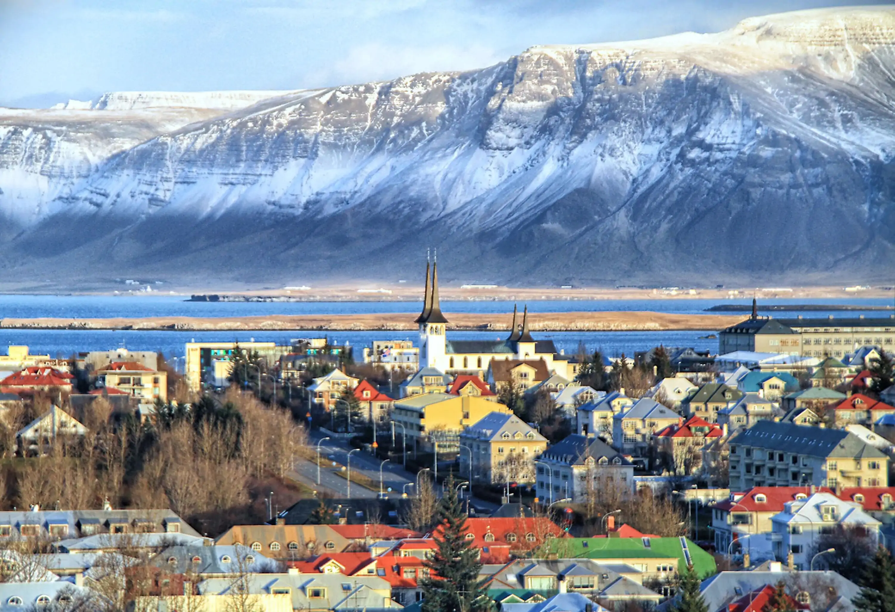 A church with twin spires overlooks a cluster of residential buildings with distant views of snow-capped mountains.