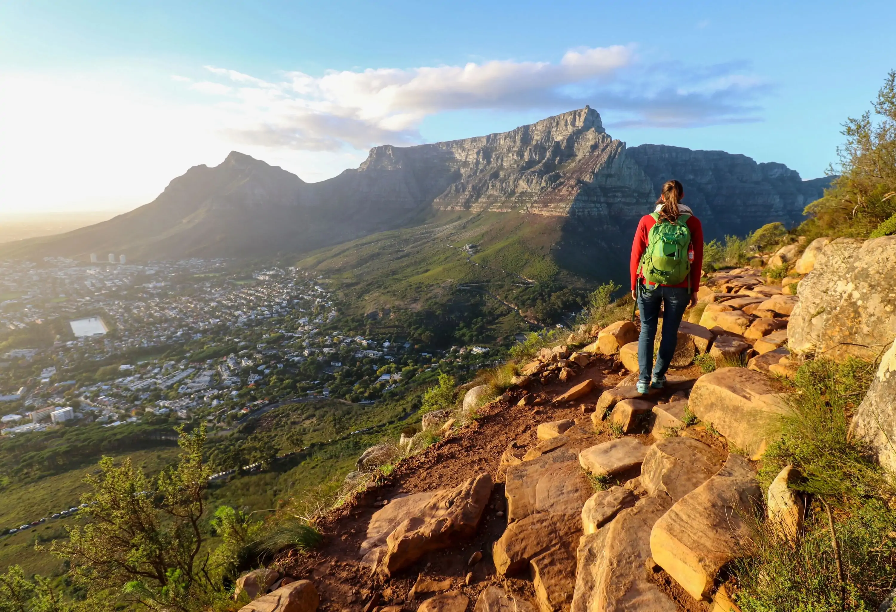 A female hiker walks on a mountaintop's rocky edge with a scenic view of a steep rock mountain over a village.