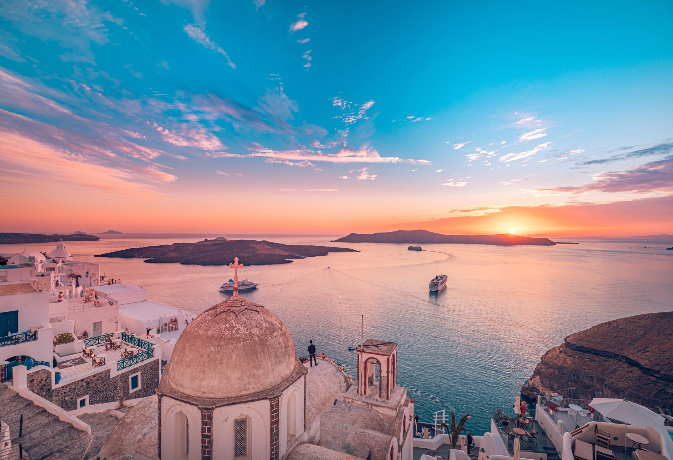 As the sun sets over the bay, the dramatic cloudy sky provides a stunning backdrop to a row of charming white stone houses and a dome-topped building, while majestic cruise ships bask in the warm glow.