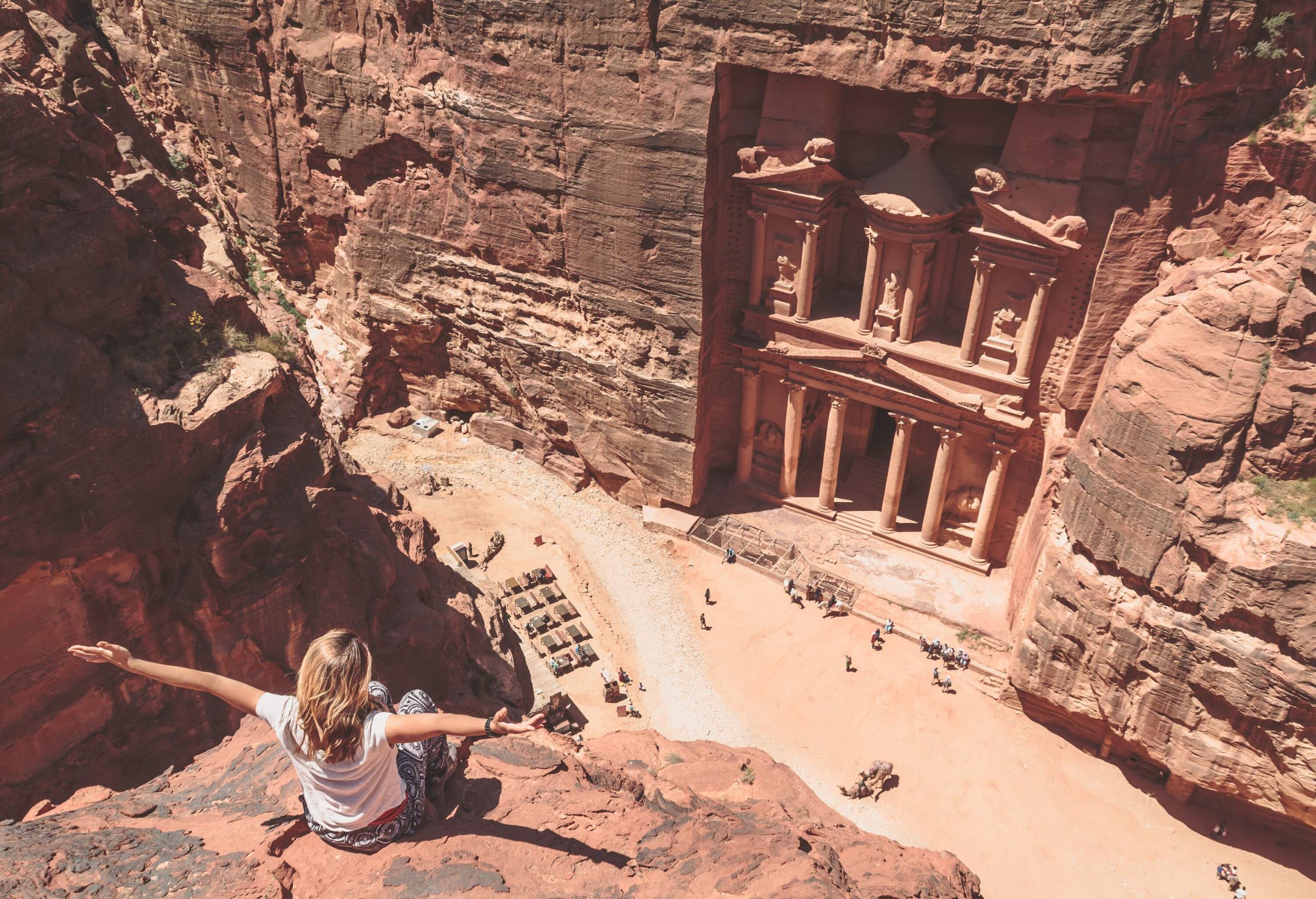 A blonde woman raised her hands sideward while sitting atop a red rock cliff facing a ruined temple carved on a sandstone rock face.