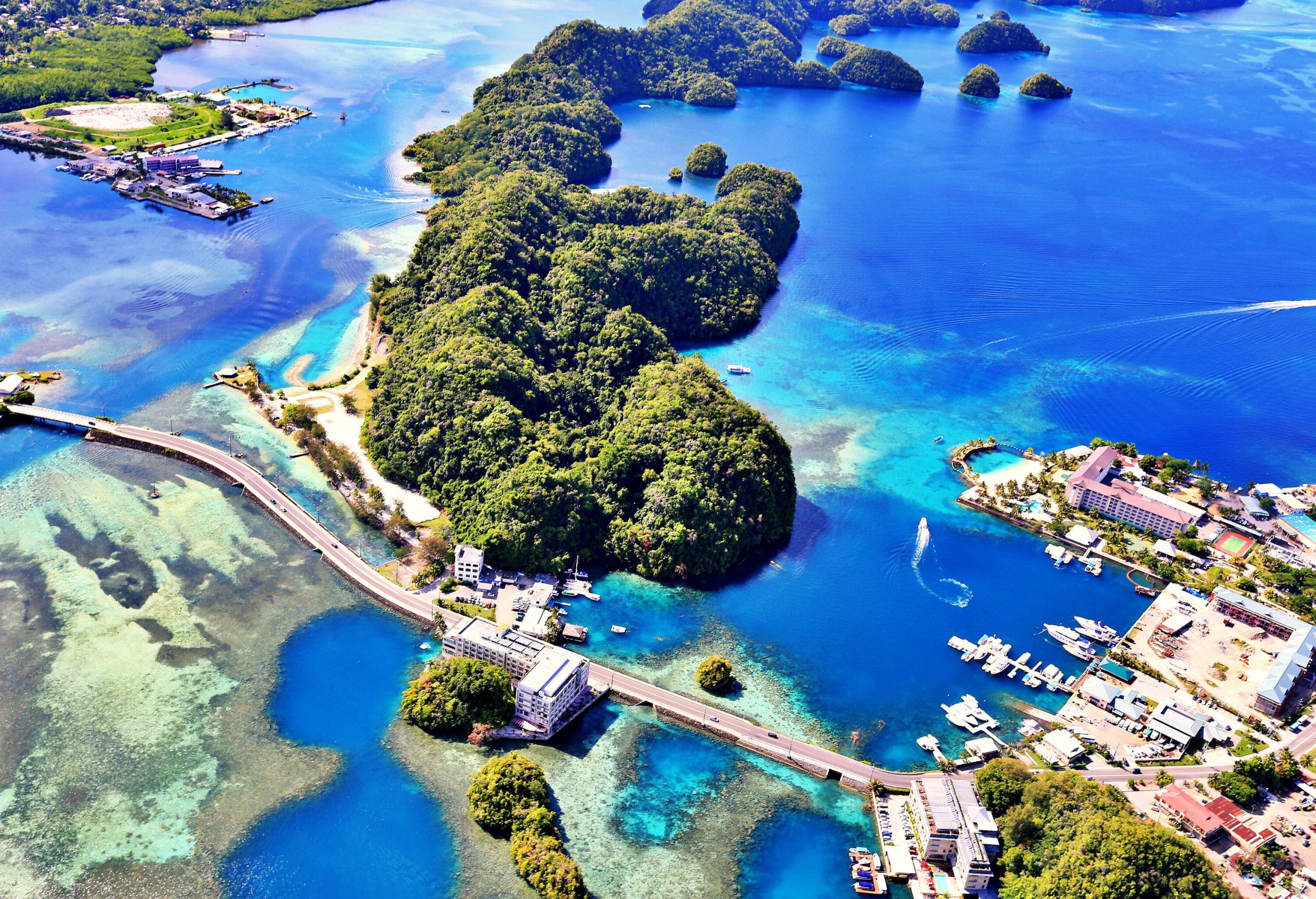 A deep blue ocean crossed by a long road bridge, a forested island, with tall buildings and docks around the coast.