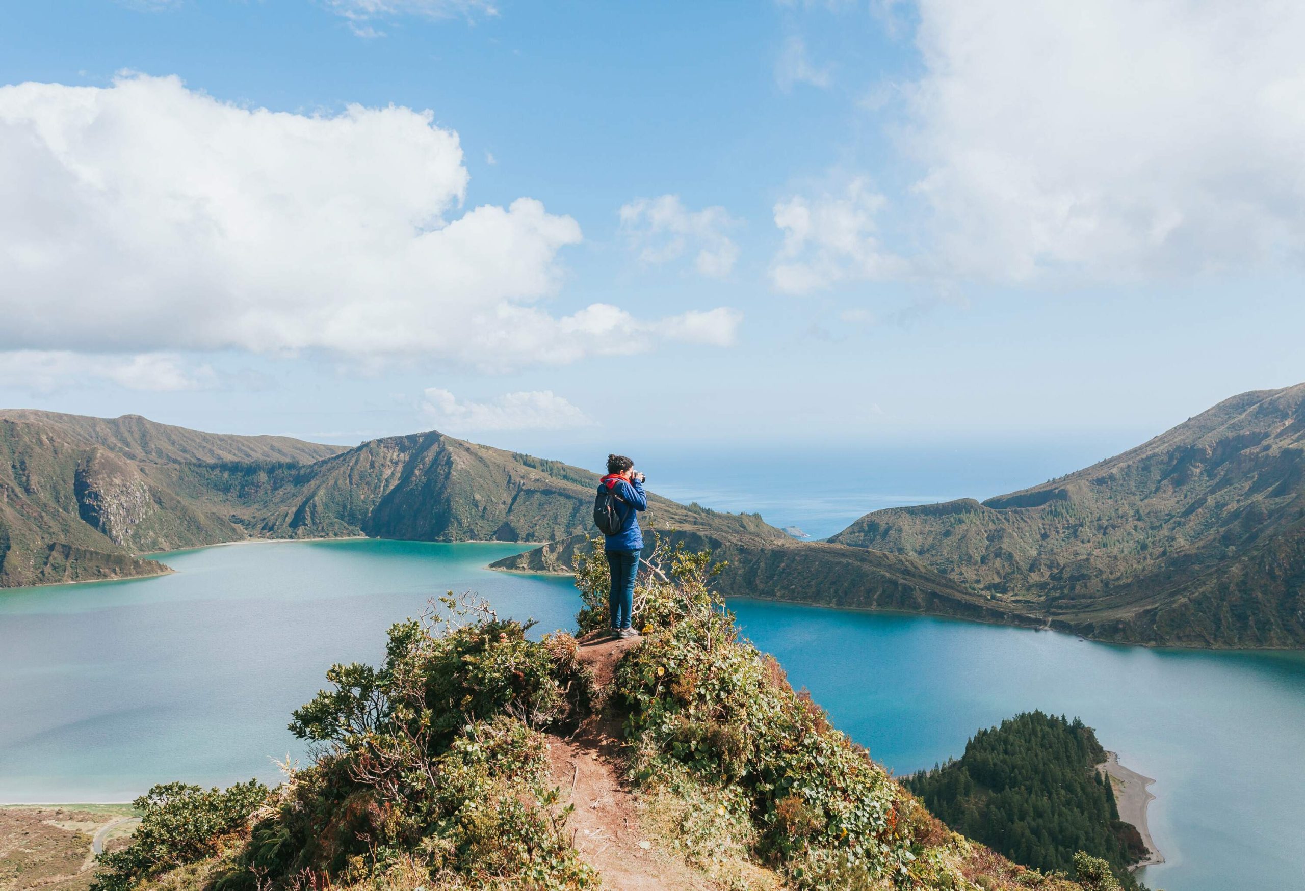 A person stands on a narrow cliff to take a photo of a turquoise lake surrounded by mountains.