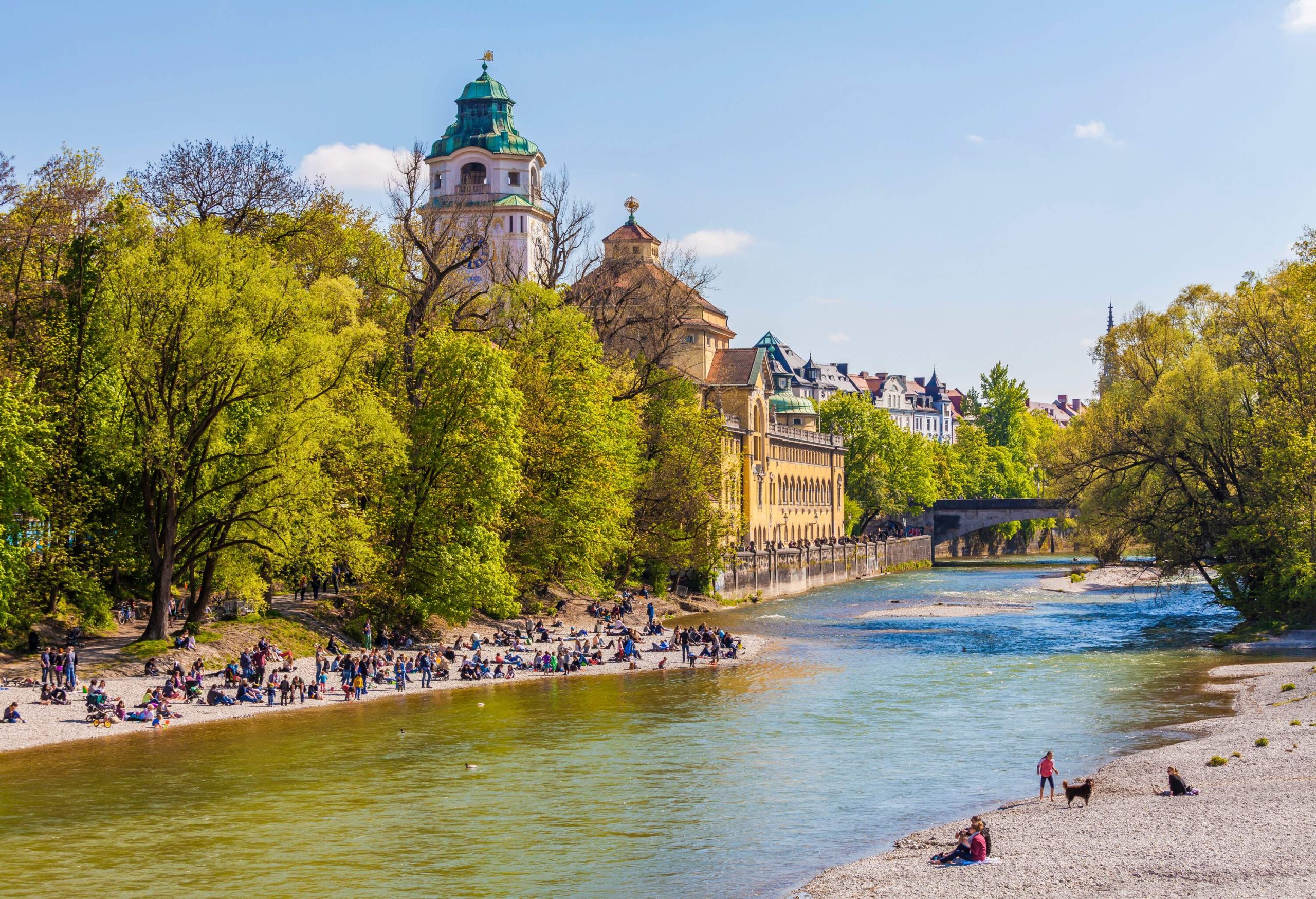 A crowd basking on the shore of a river lined with tall green trees and classic buildings.