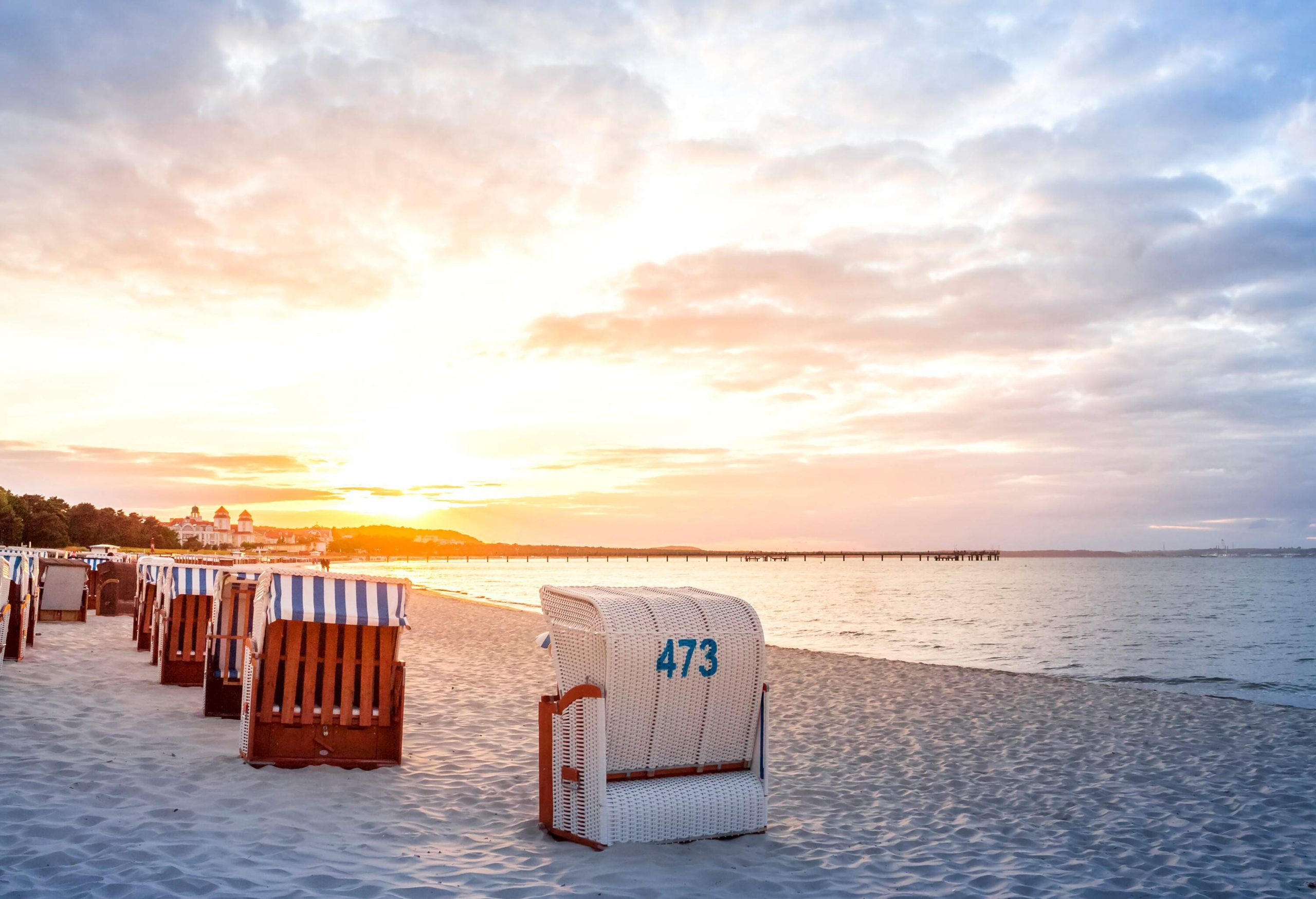 A row of roofed, wicker and wood beach chairs on the white sand shore of a beach against the dramatic sunset sky.