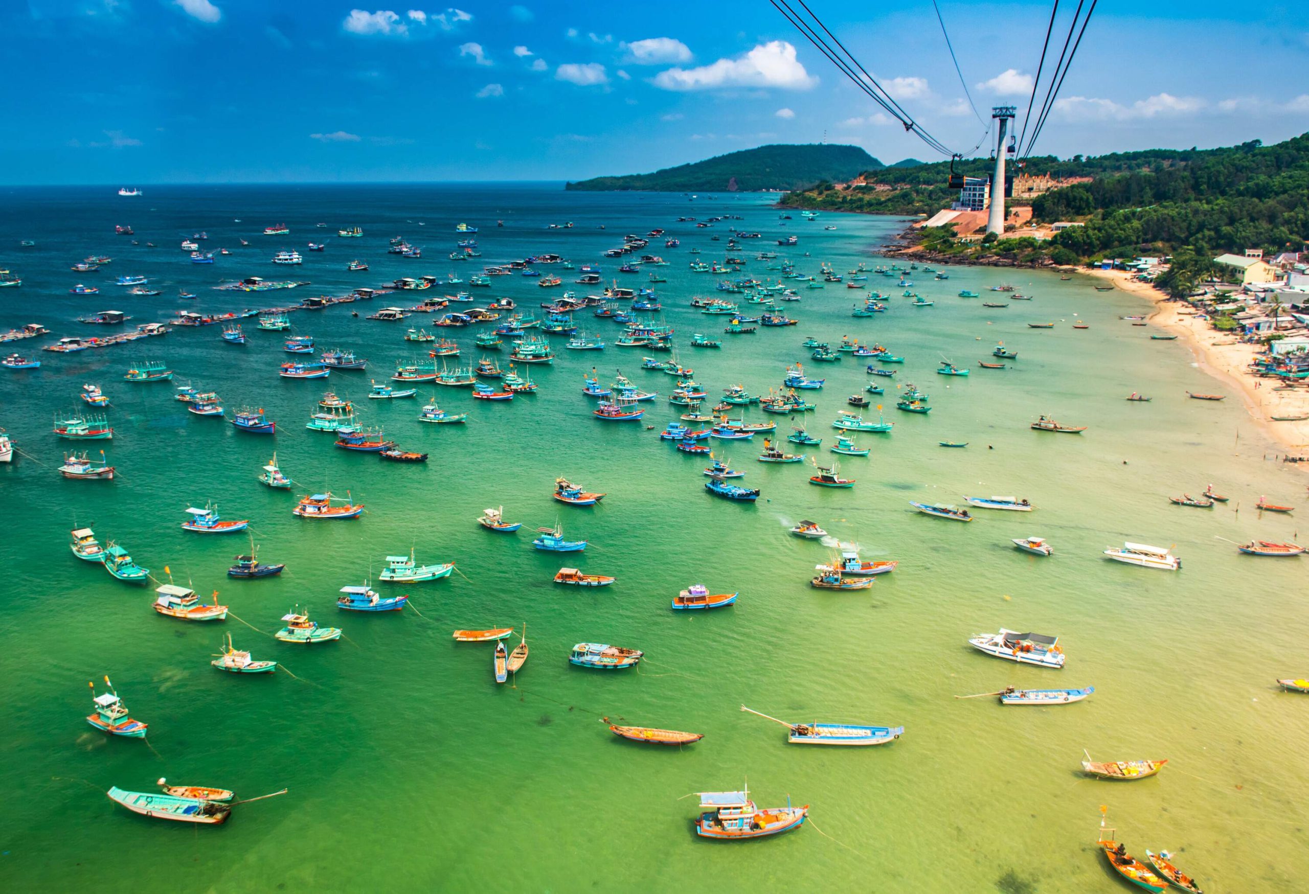 Cable cars travelling above the sea near the shore, occupied by numerous fishing boats docked on the shallow emerald green waters.