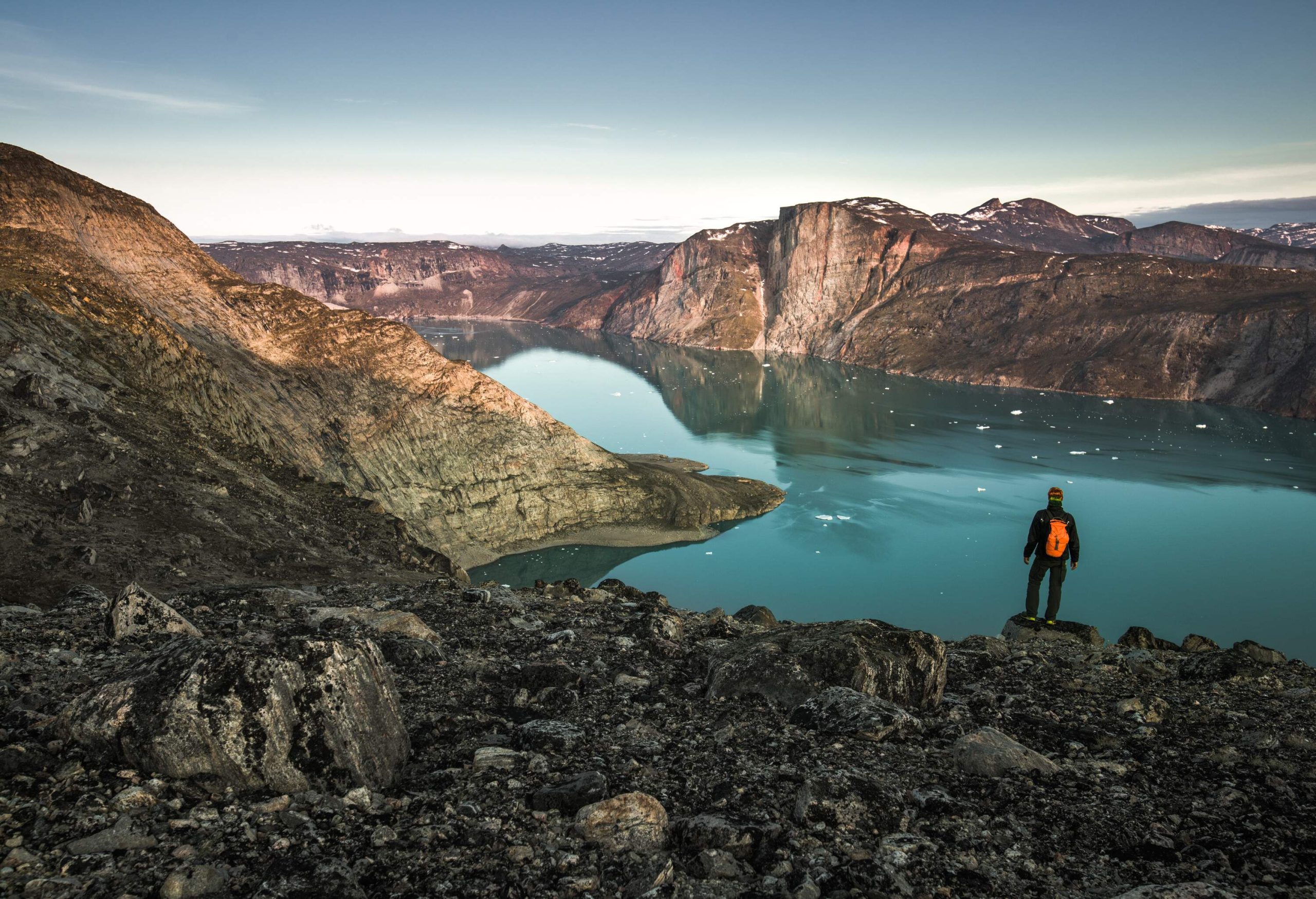 A hiker stands on a rock at the edge of a rugged mountaintop overlooking the lake enclosed by cliffs.