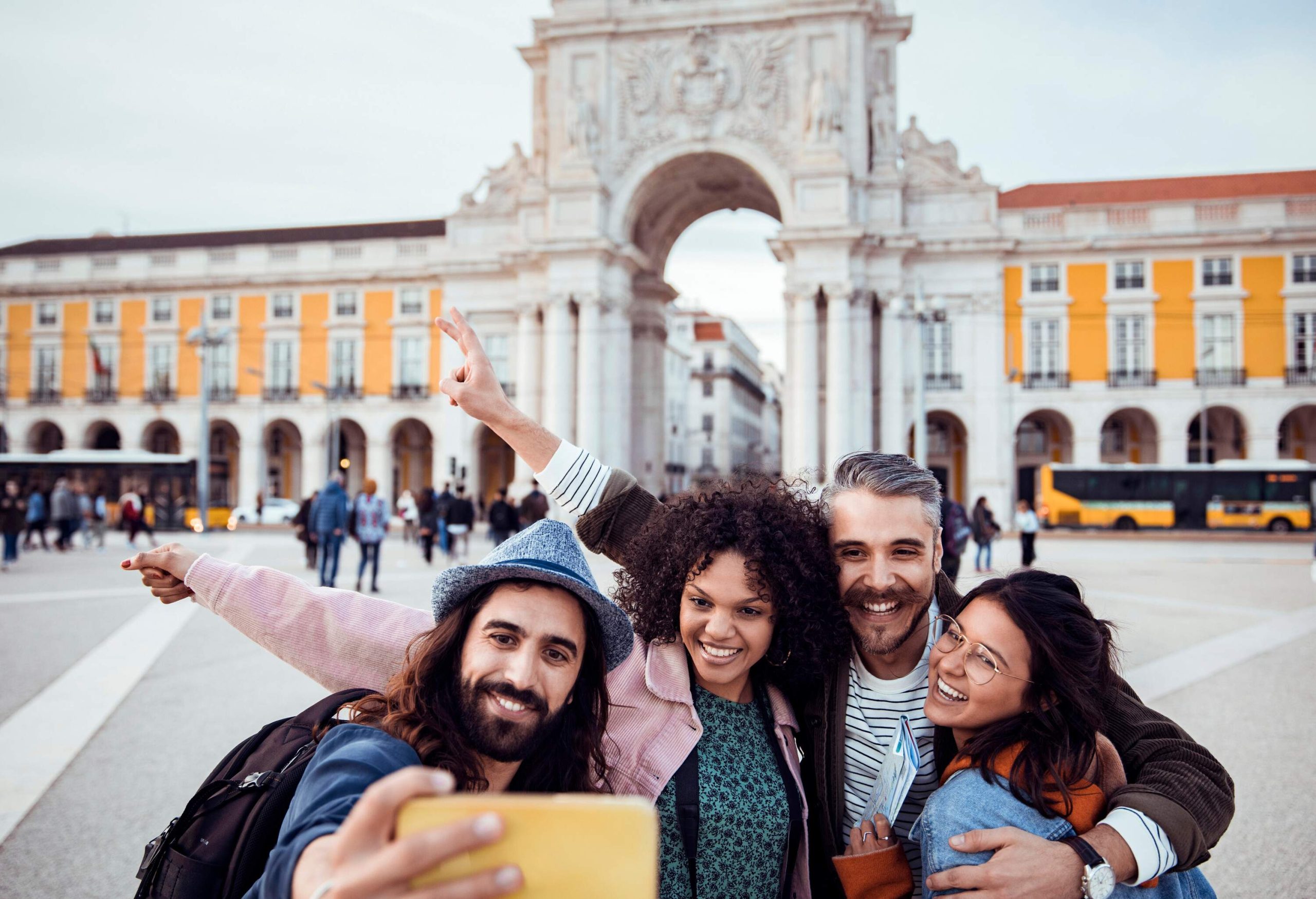 Four friends smile as they took a group photo using a smartphone in a public square with an arch monument in the background.
