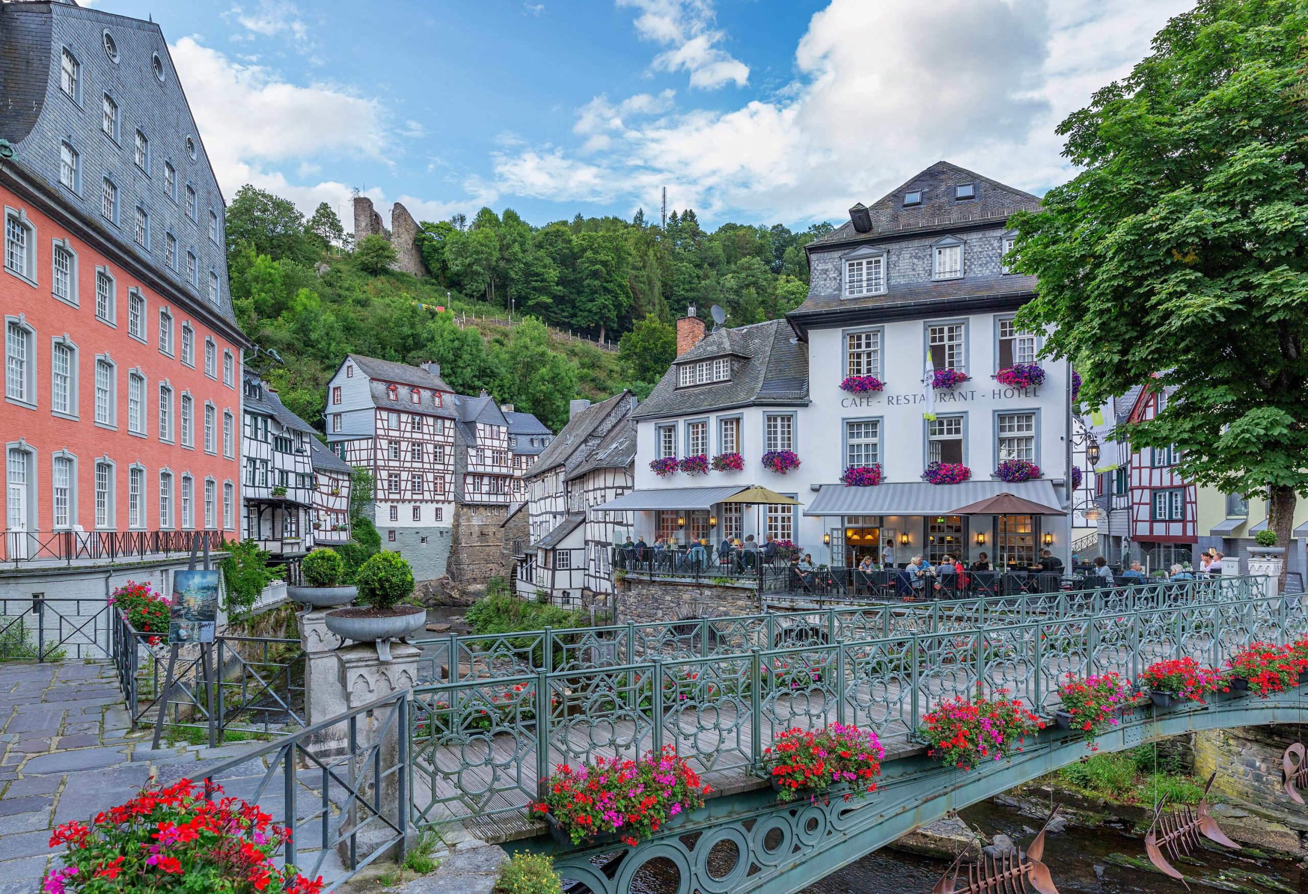 A green metal pedestrian bridge ornamented with colourful flowers across a river in the middle of half-timbered buildings.