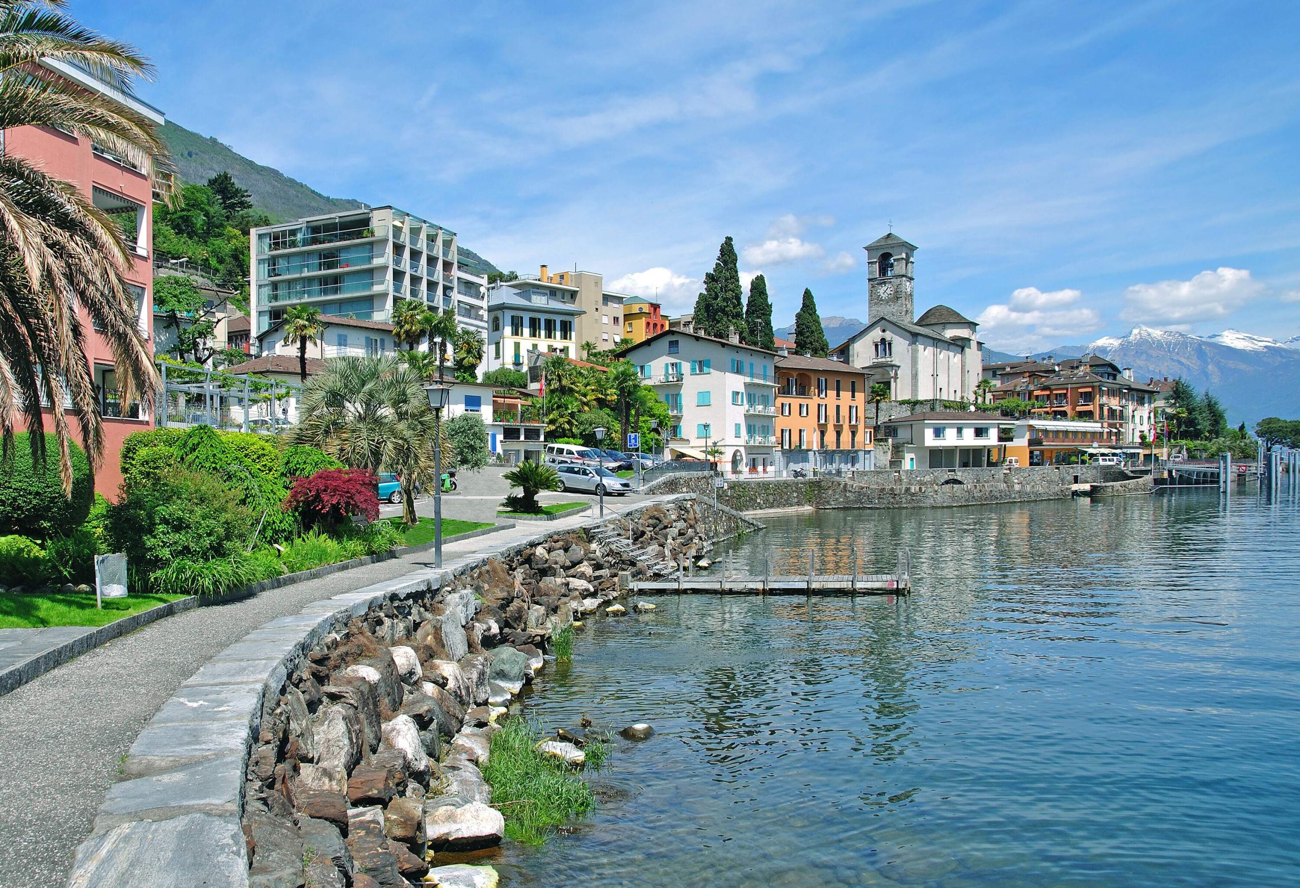 A promenade beside colourfully painted houses alongside a clear, tranquil lake.