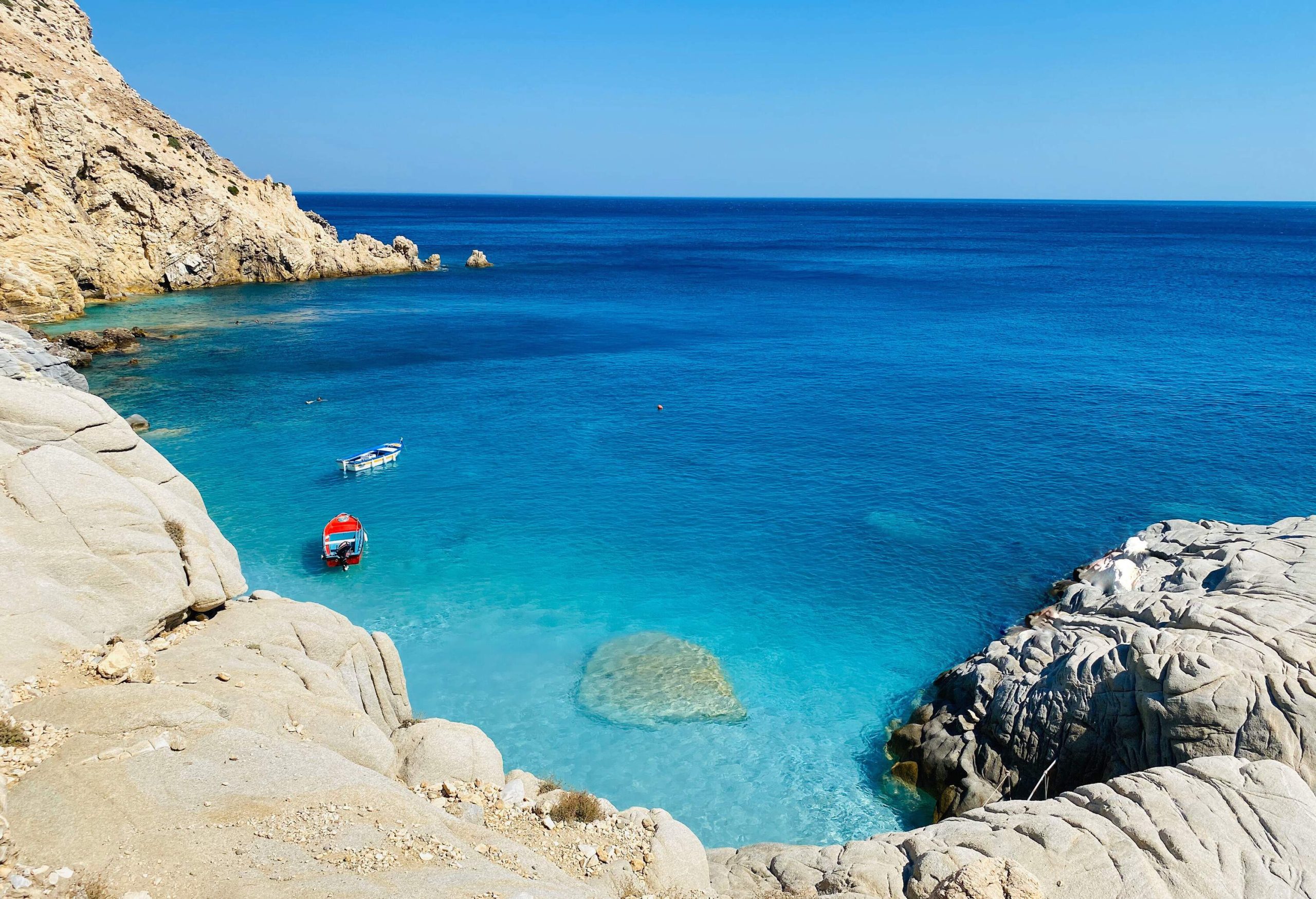A breathtaking crystal clear turquoise sea with red and white boats moored alongside the rugged rocky coast.