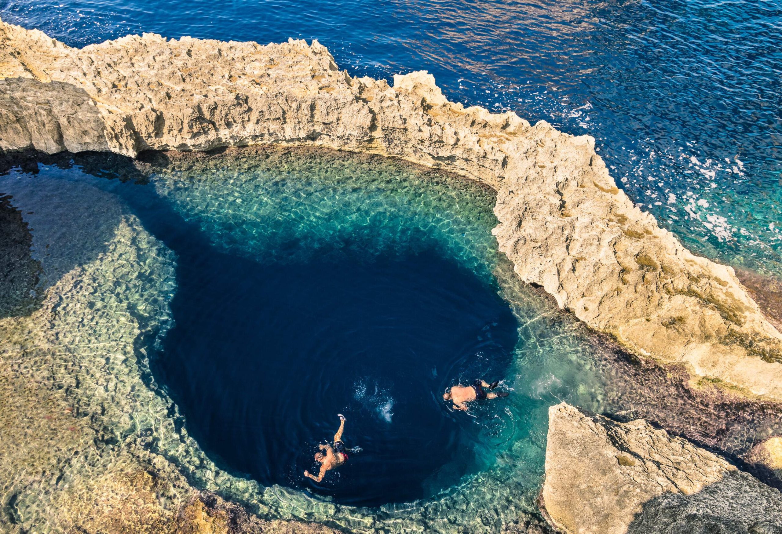 Two people are swimming in a large, deep blue hole next to the sea, which is encircled by a sedimentary rock formation.