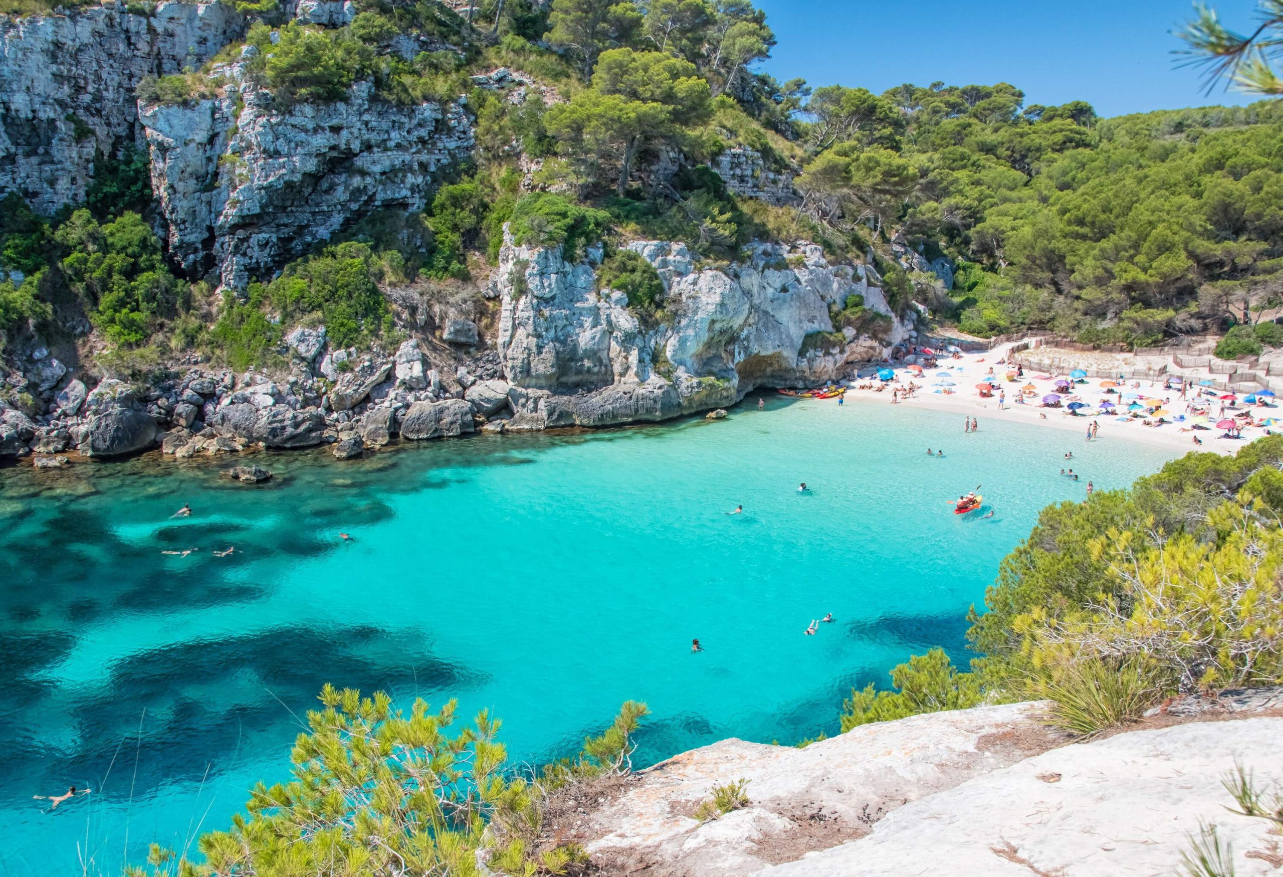 A narrow cove with people swimming in turquoise waters and relaxing on white sand.